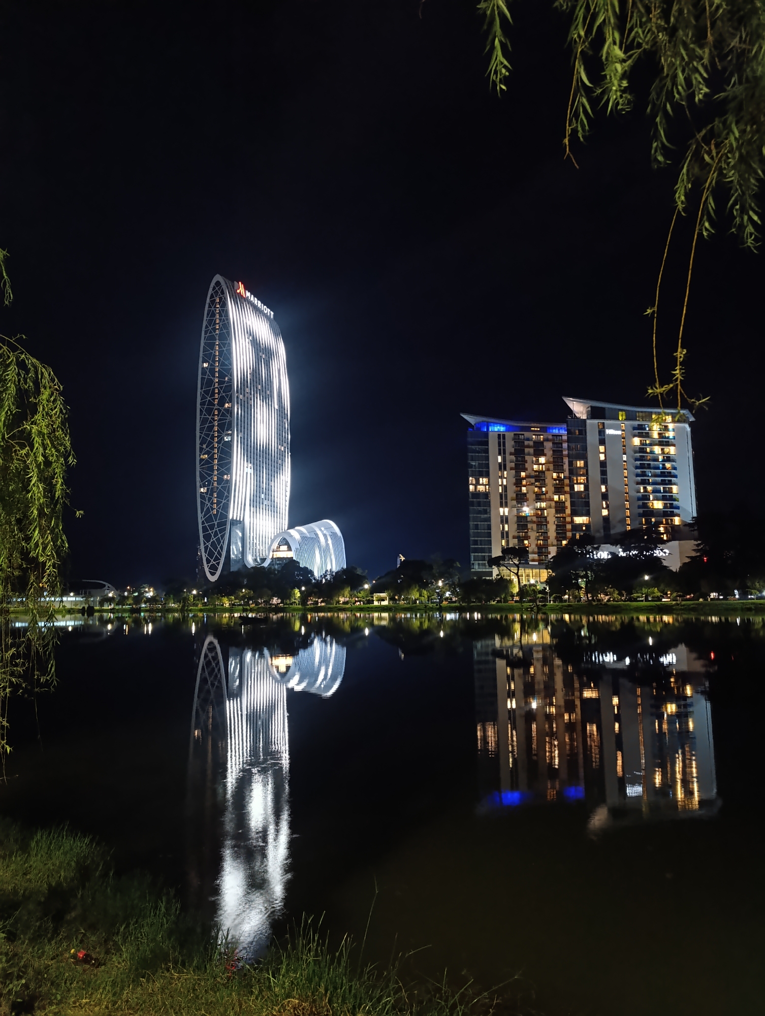 This is a nighttime, horizontal, full-frame photograph of the Batumi Boulevard in Batumi, Georgia. The image captures the unique architecture of the Marriott Batumi hotel, a tall, oval-shaped skyscraper illuminated with white vertical lights, and a rectangular, modern hotel with a blue-lit roof, reflected in the calm waters of a lake in the foreground. Trees and vegetation frame the scene on the left and right, with their reflections also visible in the water. The grass on the embankment is visible in the lower part of the frame, and there are a couple of plastic bottles on the grass. The sky is dark and clear.
