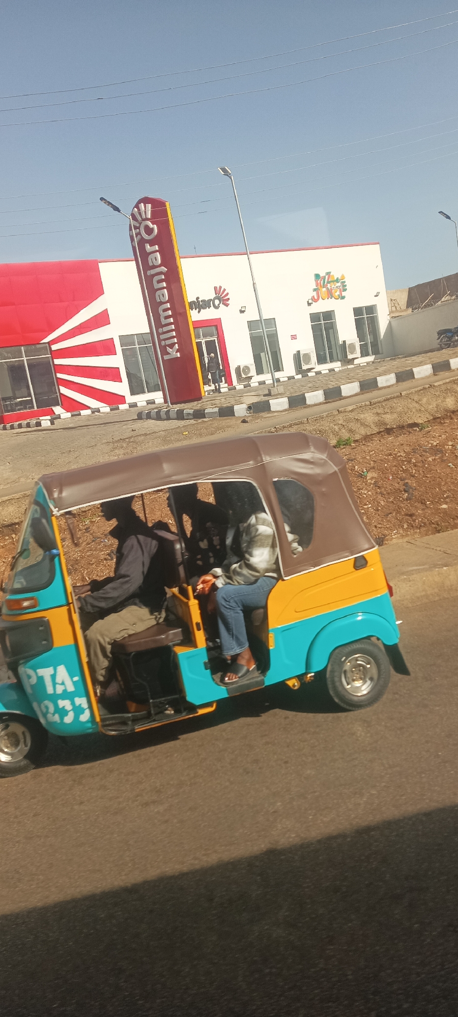 A vibrant street scene unfolds, likely in Jos, Nigeria, featuring a three-wheeled auto-rickshaw, commonly known as a keke napep, transporting passengers. The auto-rickshaw is painted in striking blue and yellow, with a brown canvas canopy providing shade. A registration number, partially visible as "PTA-1233," is printed on its side. Inside, a driver in dark clothing is at the controls, and at least two passengers are seated in the back. One passenger is wearing blue jeans and a plaid shirt, while another's silhouette is visible.
In the background, a modern commercial building with white and red facades stands under a clear, bright blue sky, indicating a sunny day. A prominent vertical sign in red and yellow reads "Kilimanjaro" in white text. The building itself also features "Kilimanjaro" branding near an entrance. Adjacent to it, another section of the building displays "Pizza & Juice" in colorful, playful lettering, suggesting a food establishment. The road surface is asphalt, and a black and white checkered curb separates the road from the building's frontage. Overhead power lines are visible against the sky. The scene captures a typical urban commute or street activity in Nigeria.