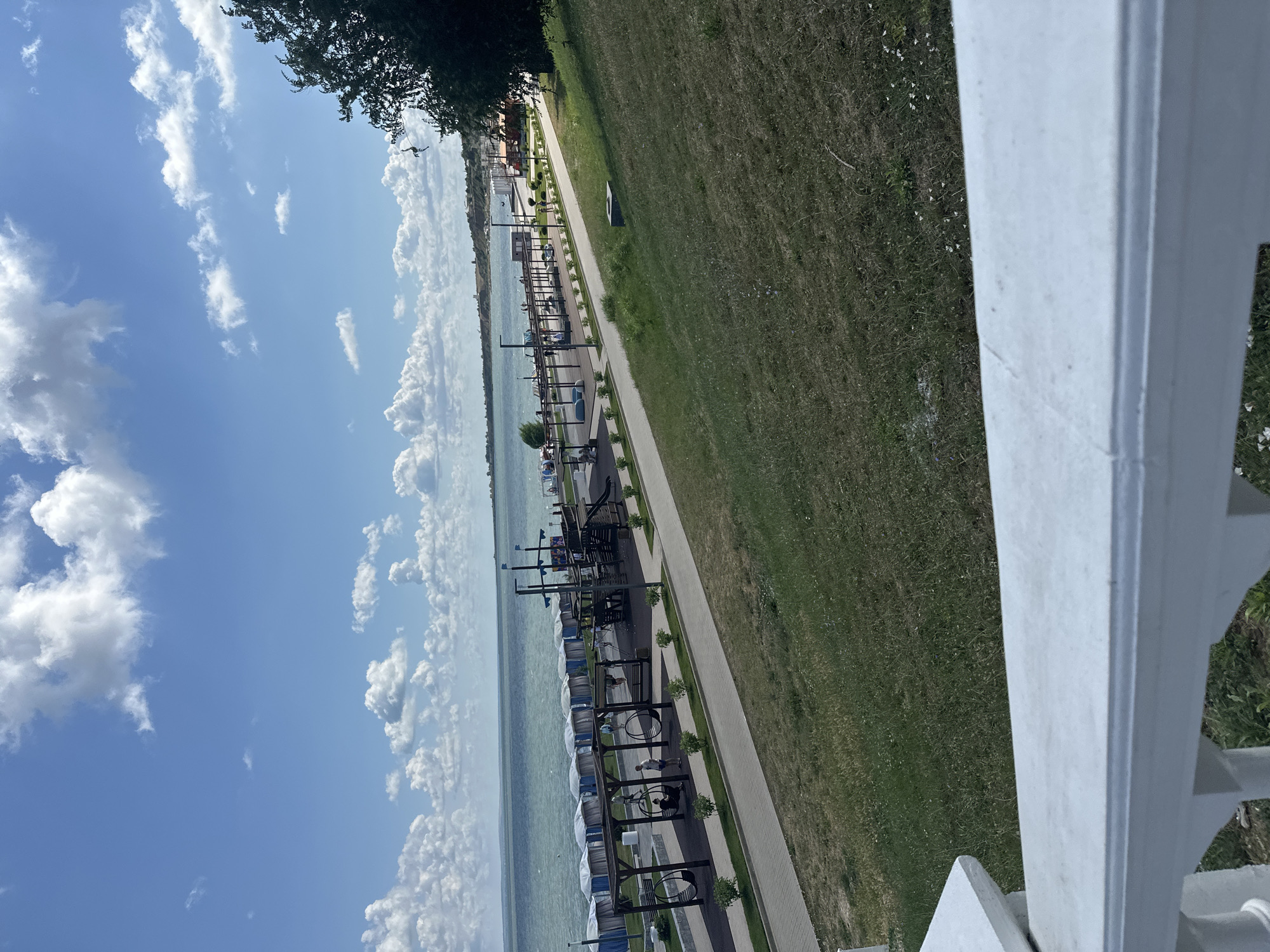 This image shows a seaside promenade in Taman, Russia, under a partly cloudy blue sky. The foreground features a white railing and a grassy slope leading down to a paved path. The path runs parallel to the sea and is lined with benches, playground equipment, and covered seating areas. People are visible walking and sitting, some near swings. Beyond the path is the sea, with calm, greenish water. In the distance, the shoreline is dotted with buildings and trees, and a lighthouse or tower is visible on a distant hill. A large tree dominates the right side of the image, obscuring part of the distant view.