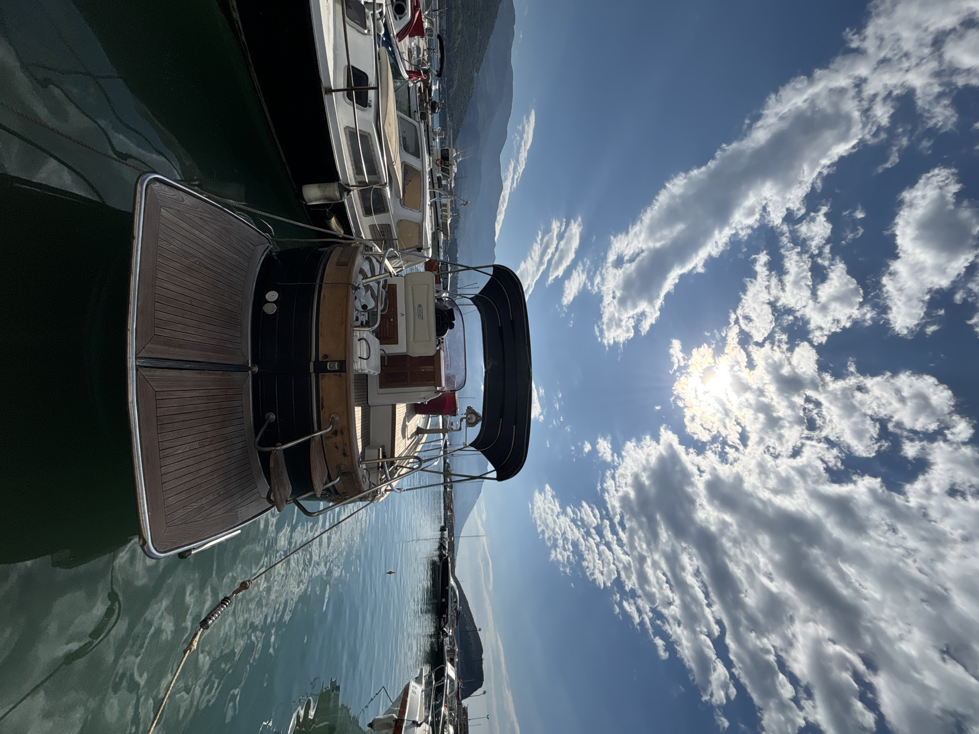 The image shows a marina in Budva, Montenegro, on a sunny day with scattered clouds. Several boats are docked, with the stern of a motor yacht featuring prominently in the foreground. The yacht has a wooden deck and black hull. Mountains are visible in the background. The water is calm and reflects the sky.
