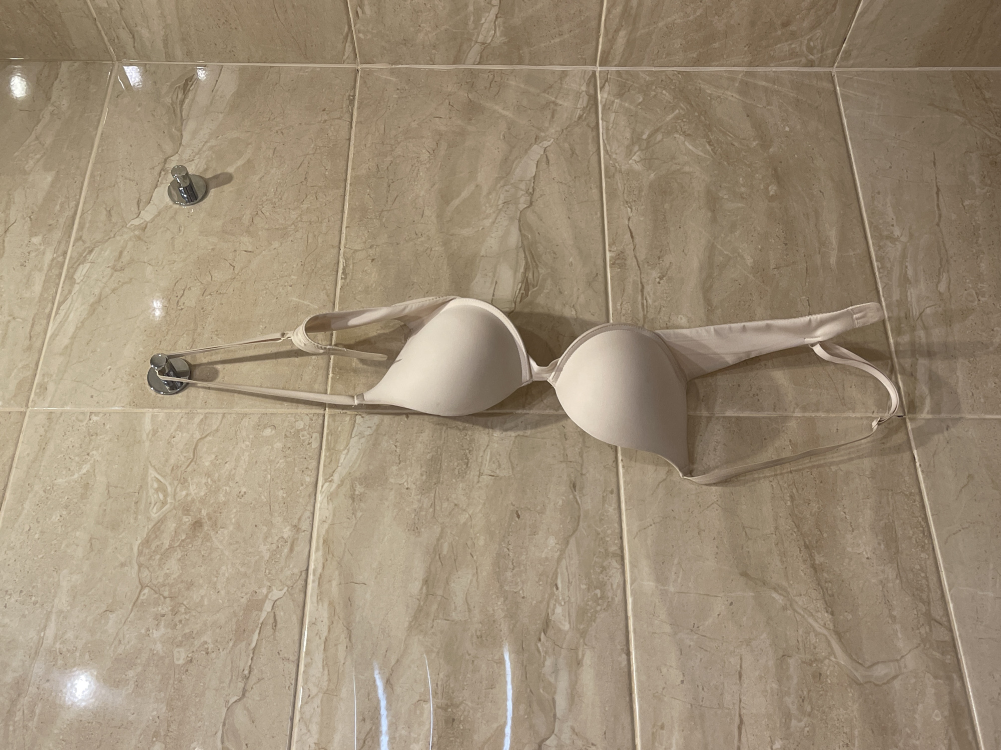 A nude-colored bra is hanging on a hook attached to a tiled wall. The tiles are beige with a marbled pattern. There are two hooks visible; one holds the bra, and another is empty. The bra is suspended by its straps, with the cups facing forward and the band hanging down. The scene appears to be in a bathroom or changing area. The location is a bathroom in a residential building in Khimki, Russia.
