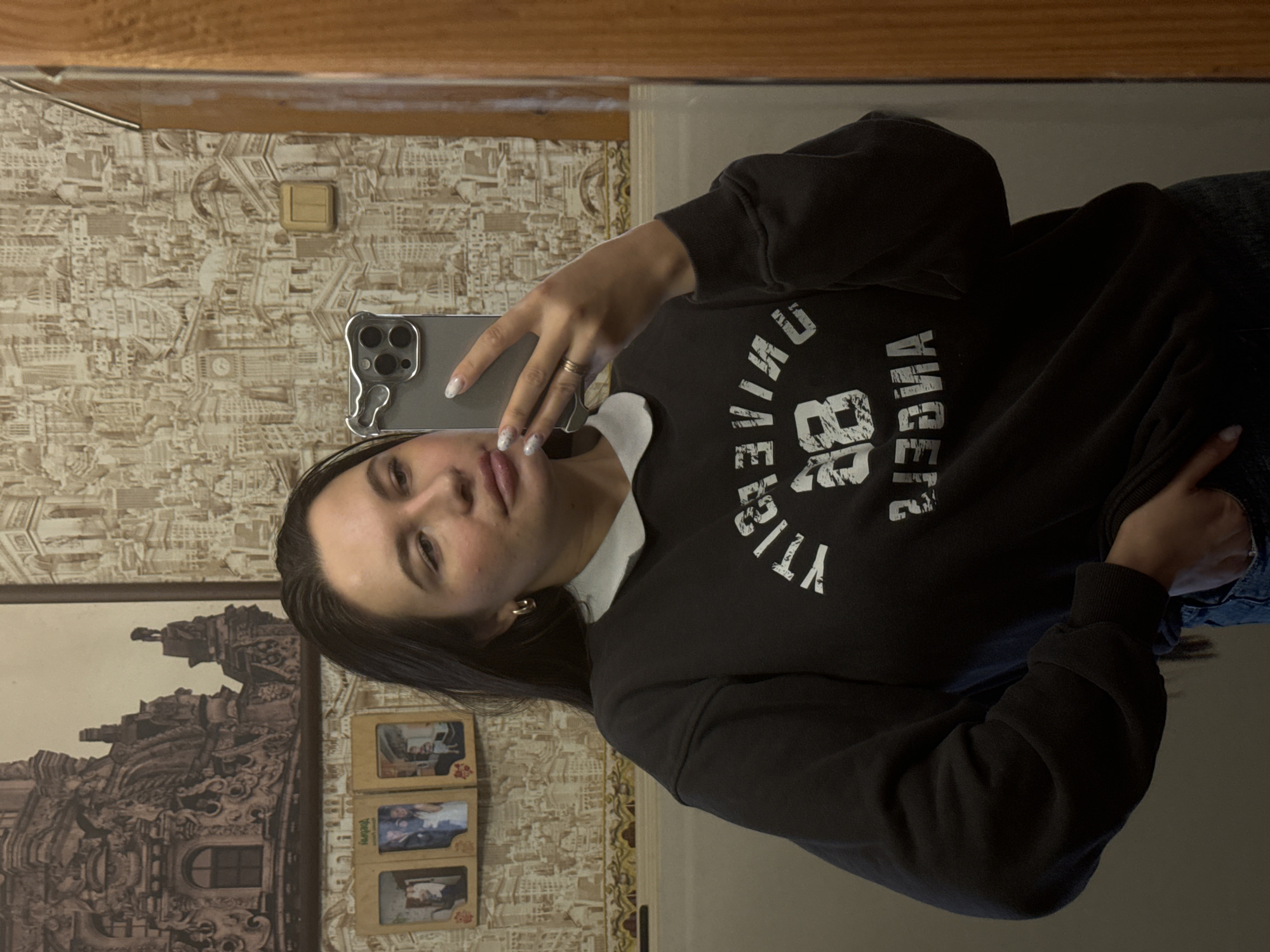 A woman with dark hair is taking a selfie in a mirror. She is wearing a black sweatshirt with white lettering. The background includes patterned wallpaper, a framed picture, and what appears to be a light switch. The location is potentially a private residence, but no definitive evidence indicates it is in Красный Маяк, Russia.