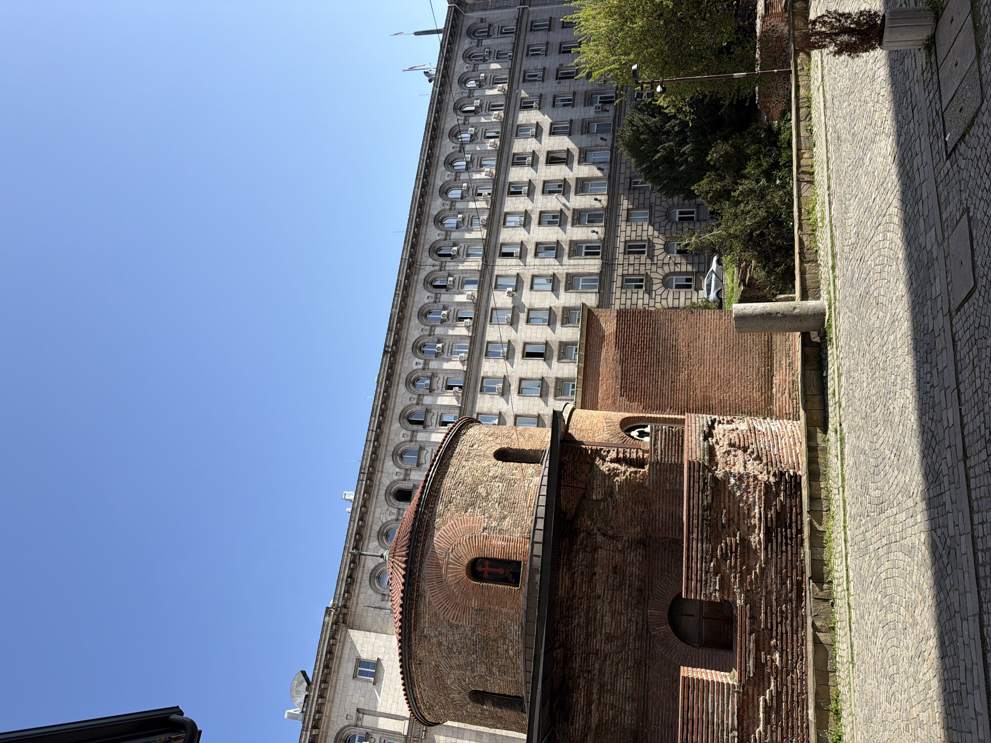 The image captures a vibrant urban scene in Sofia, Bulgaria, showcasing a striking juxtaposition of ancient and modern architecture under a clear, bright blue sky.
In the foreground and central-right, an ancient round church or rotunda, constructed from red-brown bricks, dominates the view. Its circular apse features a tiled roof and intricate brick patterns around small, arched windows. One window contains a dark panel with a prominent red cross. The brickwork displays varying textures and states of preservation, indicating its age and history.
Behind and to the left of this historic structure stands a much larger, multi-story building with a light-colored facade, likely stone or granite. This modern building features several rows of windows; the upper visible floors have arched windows, while lower ones are rectangular. Numerous white air conditioning units are attached below many of the windows. Flags are visible flying from flagpoles on the building's roof. Green trees and bushes partially obscure the left side of this building and are also visible near the ancient structure.
To the right of the brick building, a well-maintained cobbled street or pedestrian area with a distinctive arc pattern stretches into the background. Strong sunlight casts sharp shadows across this area, indicating a sunny day. A street lamp post and a thick stone column or base are also visible along the cobbled path.
The scene is devoid of people, and no legible text appears, aside from the symbolic red cross on the ancient church. The clear blue sky and crisp lighting suggest it is daytime, likely morning or early afternoon. The overall impression is one of historical depth and architectural contrast, typical of central Sofia where historical layers are integrated into the modern city fabric.