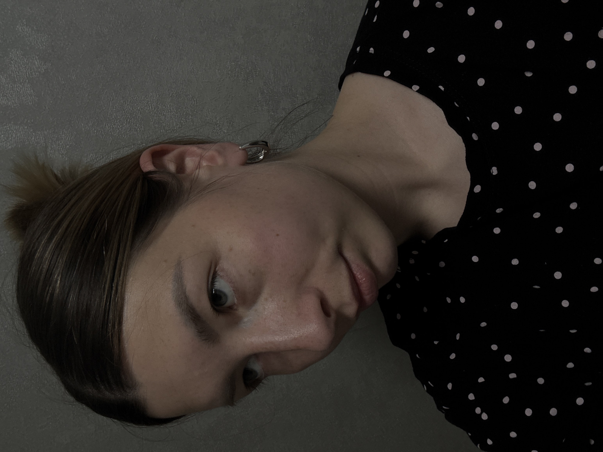 A close-up, low-angle shot presents a woman with fair skin, positioned at an oblique angle against a textured gray background. She is wearing a black top patterned with small white polka dots. Her dark blonde hair is pulled back into a loose bun, with visible highlights. She has a silver hoop earring on her left ear, and her facial features—including eyes, nose, and lips—are clearly visible. The lighting in the image is somewhat dim, creating shadows on her face and neck. There is no information that indicates this image was taken in Moscow, Russia.