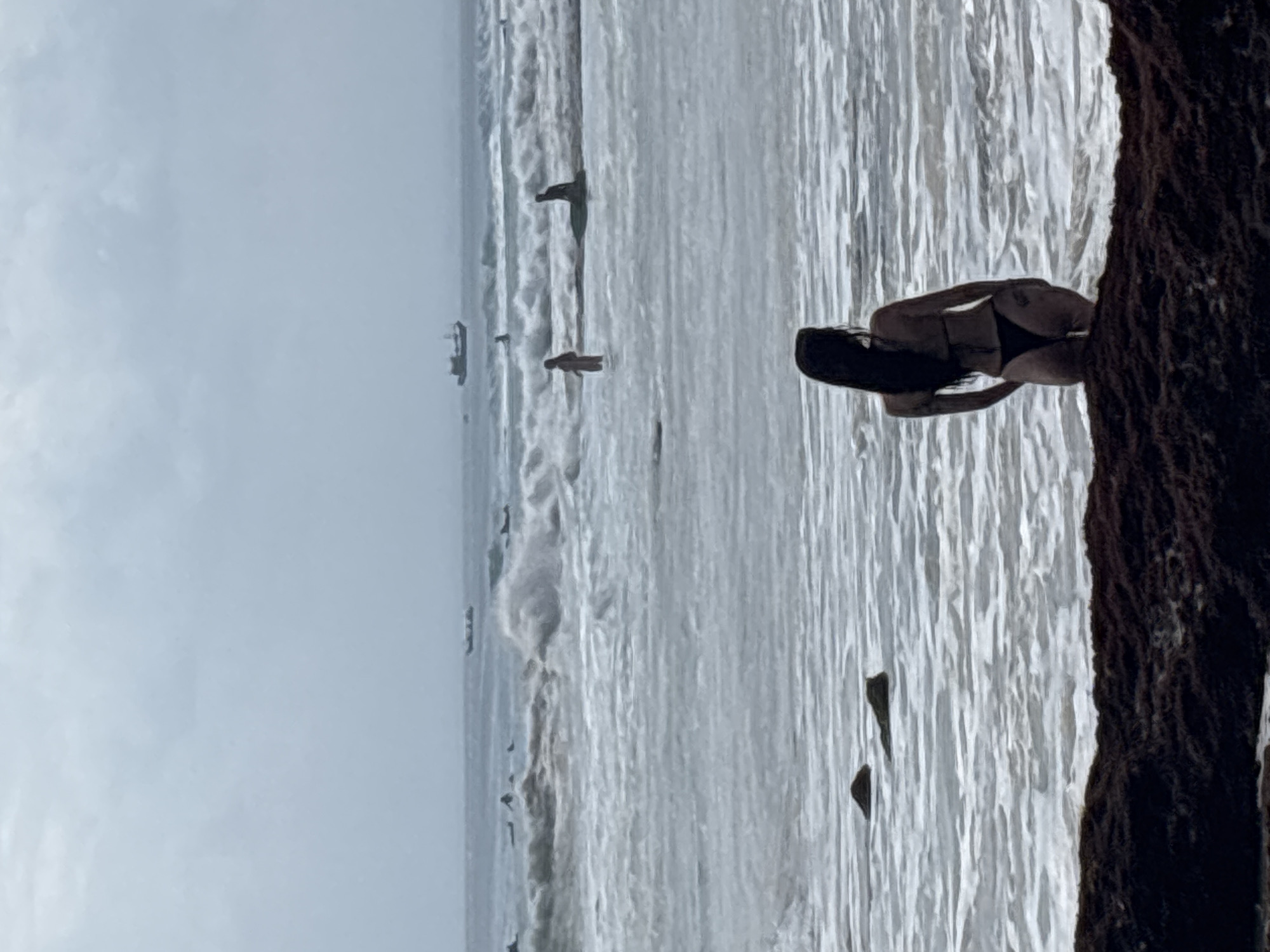 A woman in a black bikini stands in shallow water on a rocky shore, facing away from the camera towards the ocean. Her long, dark hair is down her back. In the distance, the sea is rough with large waves. Several people are visible in the water, some appearing to be surfers. Two boats are also in the distance. The sky is overcast. The scene is likely the coastline of Kuta Selatan, Indonesia, known for its surfing beaches and rocky shores.