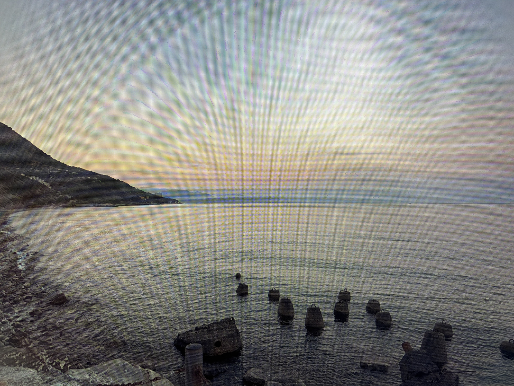 A coastal scene at sunrise or sunset depicts a calm body of water with a rocky shoreline. Several dark, cylindrical objects with visible loops are submerged in the water, partially breaking the surface. A landmass with vegetation and structures is visible in the background, sloping towards the water. The sky is a gradient of pale yellow, pink, and blue, with a visible lensing effect across the top portion of the image. The scene is devoid of any human figures. The location is the city of Mirnoye, Ukraine.