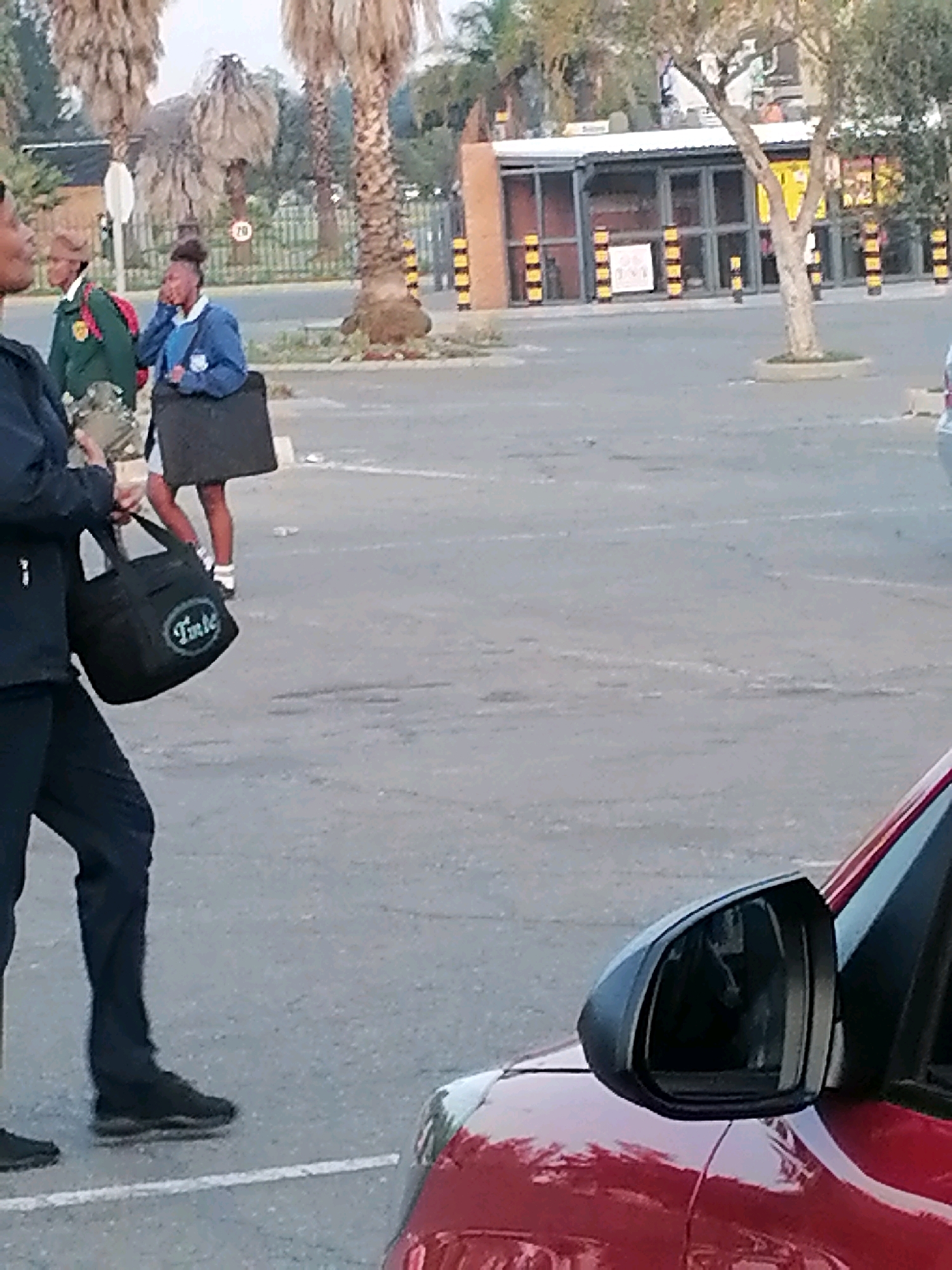 The image depicts an outdoor scene in Emfuleni Local Municipality, South Africa. In the foreground, a person in dark clothing and black shoes is walking on a paved surface. To the left, a black tote bag with the word "Tinte" in a white oval logo is visible. In the background, two young individuals, one in a blue school uniform and the other in a green and red uniform, are standing. The person in the blue uniform is carrying a black satchel. A palm tree and other vegetation are present. Buildings and a fence can be seen in the distance, suggesting a public or school area. A red vehicle is partially visible in the lower right corner of the frame, with its side mirror in view. The lighting suggests it is daytime.