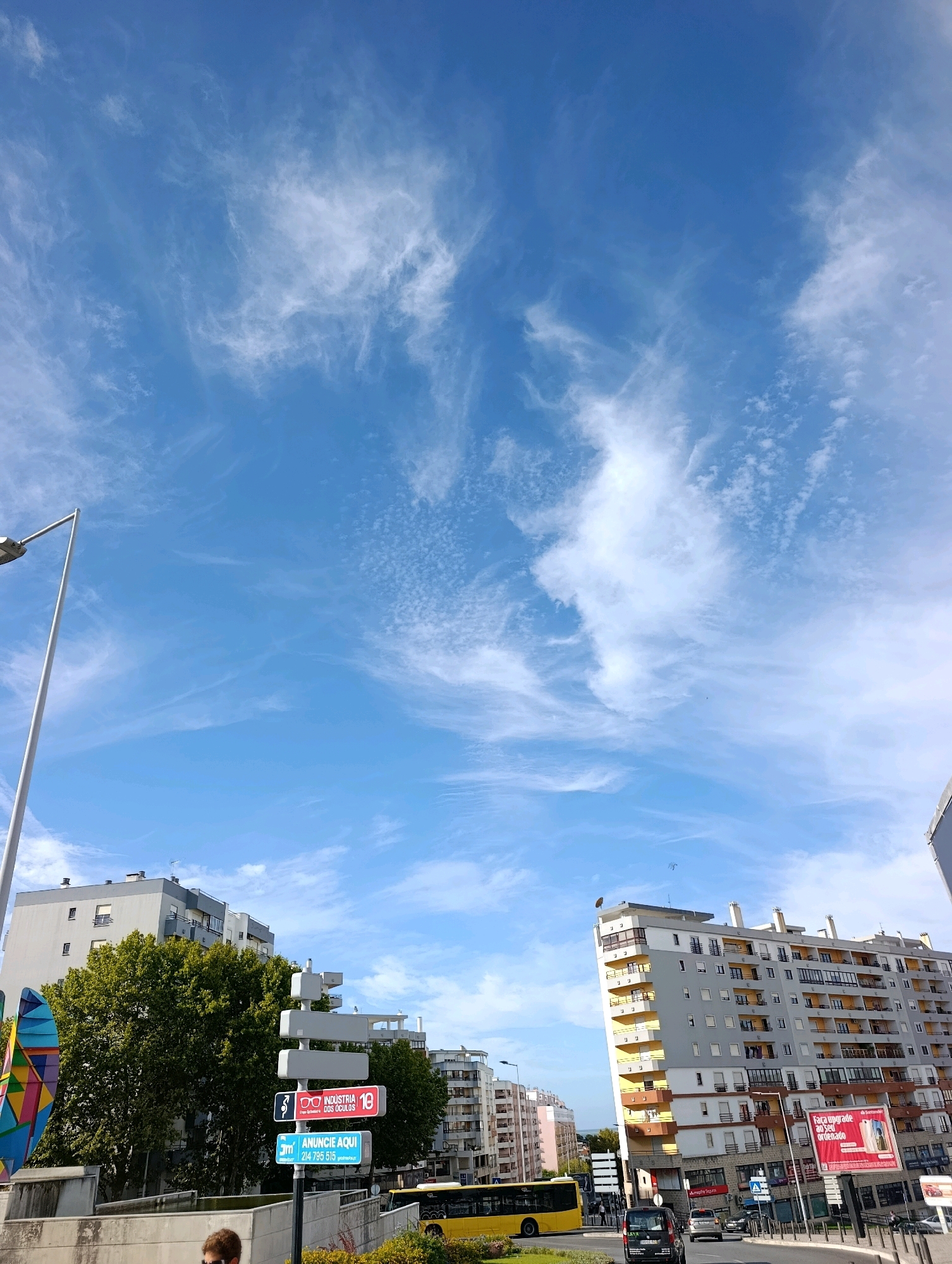 The image captures a street scene in Setúbal, Portugal, on a bright, clear day with wispy clouds in a vibrant blue sky. In the foreground, a street sign with multiple directional panels is visible. One panel reads "INDUSTRIA DOS OCULOS" with a red border and eyeglasses icon. Another below it says "ANUNCIE AQUI" with a blue border and phone number. To the left of the sign, a portion of a colorful, abstract sculpture is present. Further back, a yellow bus is driving on the street, alongside other vehicles and a black van. Tall, multi-story apartment buildings line the street, some with balconies and apartment windows. A street lamp with a white pole stands on the left side of the frame. Dense green trees are situated between the buildings and the street. The overall scene depicts a typical urban environment in Setúbal, with residential buildings, traffic, and public amenities under a wide-open sky.