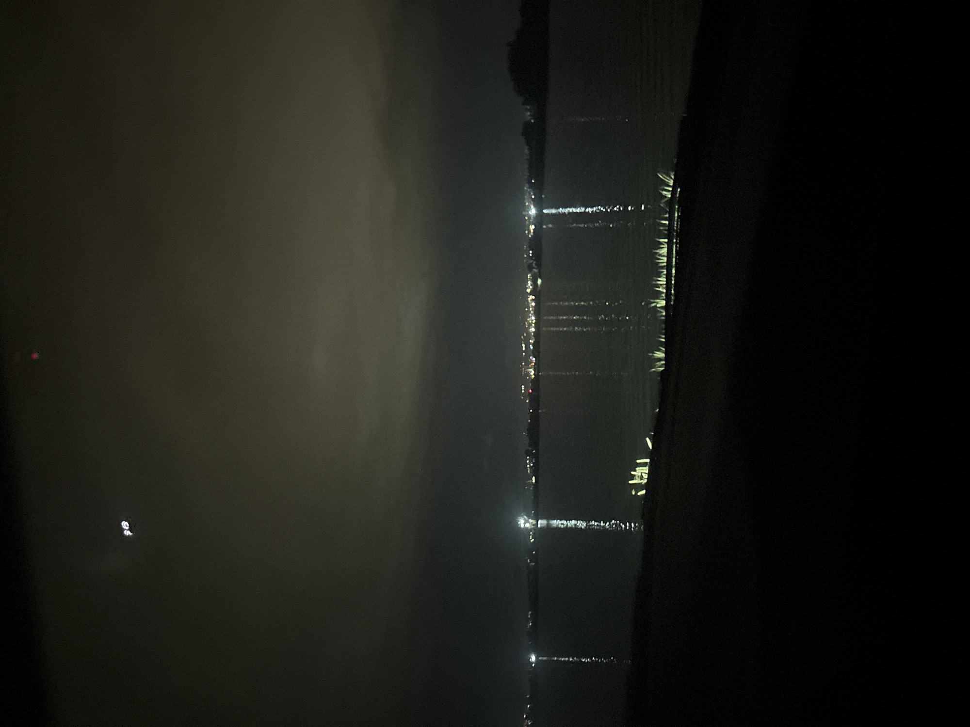 The image captures a dark, nocturnal scene of a body of water, likely a river, lake, or bay, with a distant shoreline under an overcast or hazy sky. The original image is in portrait orientation but depicts a landscape view, suggesting it was captured with the camera rotated 90 degrees counter-clockwise.
The primary features are numerous artificial lights from what appears to be a distant settlement or city on the far bank. These lights cast long, shimmering reflections onto the dark, calm water, creating vertical streaks of white and yellow across the surface. Some smaller, fainter lights are also visible closer to the foreground.
The sky, occupying the upper portion of the frame (or the left side in the original orientation), is uniformly dark and appears misty or hazy, obscuring any celestial bodies. A faint red glow, possibly a distant light source or reflection, is visible in the upper-left corner of the image (or mid-left in the original orientation). A small cluster of very faint white specks, possibly distant lights or noise, is also visible in the hazy sky.
The immediate foreground on the right side of the image (or bottom in the original orientation) is a dark, indistinct mass, possibly a landmass or a structure bordering the water. The overall atmosphere is quiet and serene, dominated by the darkness and the interplay of artificial lights with the water and hazy sky. No people, specific objects beyond the lights and water, or visible text are discernible.