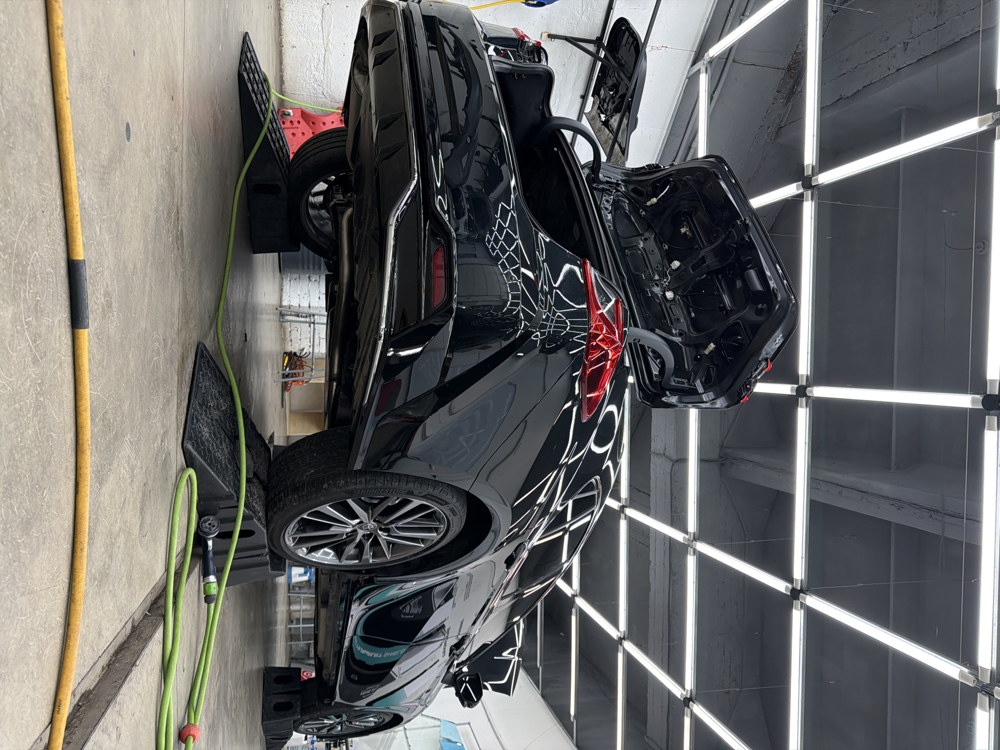 A black car, possibly a sedan or coupe, is parked in what appears to be a detailing or repair shop. The car has its driver's side door and rear trunk open. The car is elevated on black ramps. A green hose with a spray nozzle is visible next to the car on the left side. The shop is illuminated by a grid of bright white fluorescent lights. The walls and ceiling are gray. There is no indication of the city of Krasnoyarsk or Russia in this scene.