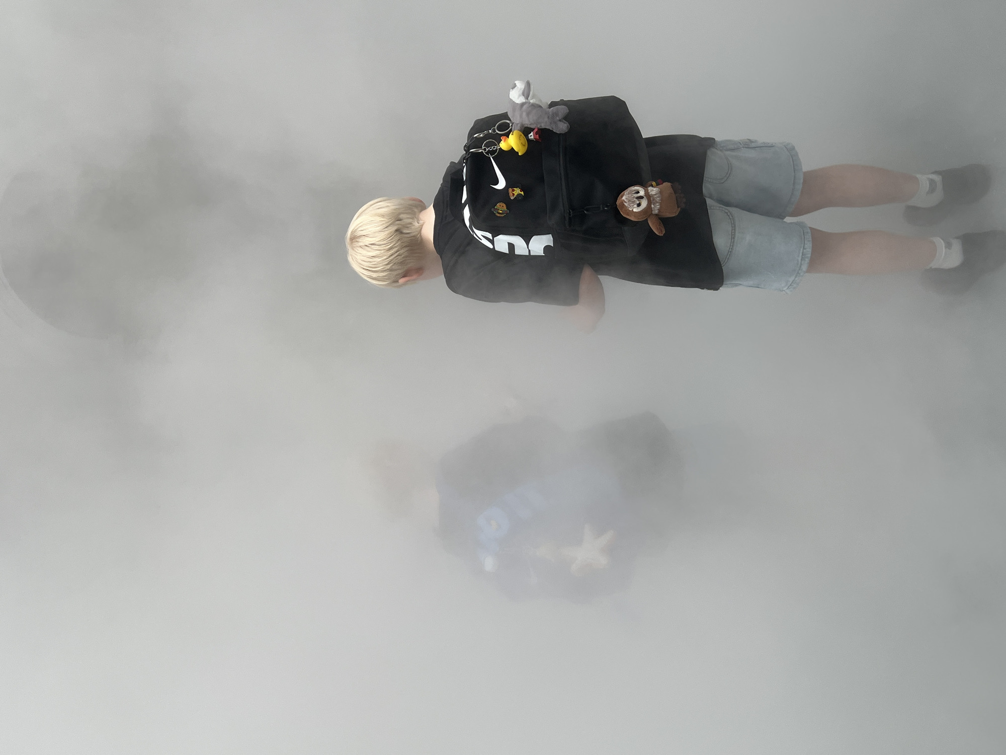 The image shows two young boys, partially obscured by fog, in what appears to be an urban outdoor environment, possibly a park or public space. The boy in the foreground, on the right, is wearing a black t-shirt with a white Nike logo and light blue denim shorts. He has blonde hair and is carrying a black backpack adorned with several keychains: a yellow rubber duck, a gray shark, and a small brown owl. The boy on the left is less visible, but appears to be wearing a dark blue t-shirt with a white graphic and a backpack. The overall atmosphere is hazy and ethereal, suggesting a foggy or misty day. The context of Paris, France, is not discernible from the visual information presented in the image.