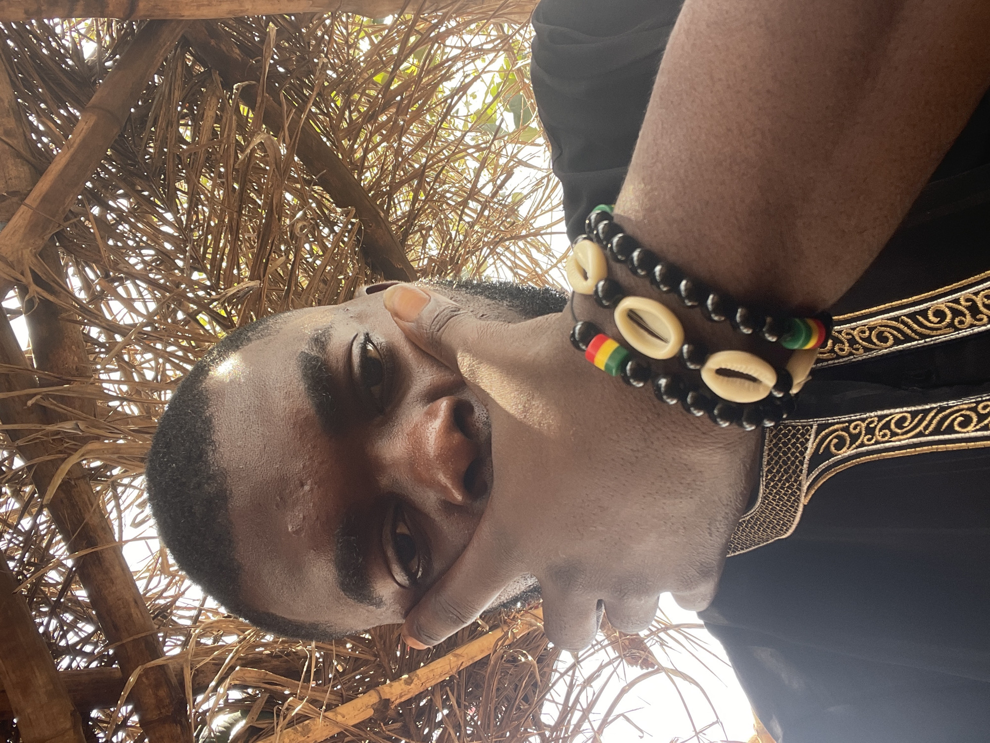 The media file presents a close-up portrait of a person with dark skin, oriented such that their head is on the right side of the frame, tilted. The camera appears to be positioned below the subject, looking upwards. The person has short, dark hair and is wearing a black garment adorned with intricate gold embroidery along what appears to be a collar or trim.
On the person's right wrist (visible on the left side of the image due to the rotation), two distinct bracelets are worn. One bracelet features black beads interspersed with light-colored cowrie shells. The second bracelet consists of black beads alongside segments of red, yellow, and green beads, often associated with Rastafarian colors. The person's left hand is raised, partially covering their chin and lower face, with their fingers lightly resting on their cheek. Their eyes are directed towards the viewer.
The background reveals a dense, dry, and interwoven structure of branches, twigs, or palm fronds, suggesting a natural canopy or a thatched roof. Bright sunlight filters through the gaps in this structure, indicating an outdoor setting during daytime. No specific weather conditions are discernible beyond the bright light. The overall scene suggests a location within Ogbe, Nigeria, consistent with the use of natural building materials and culturally significant accessories. No visible text is present in the image. The person's expression is partially obscured by their hand, but appears calm.