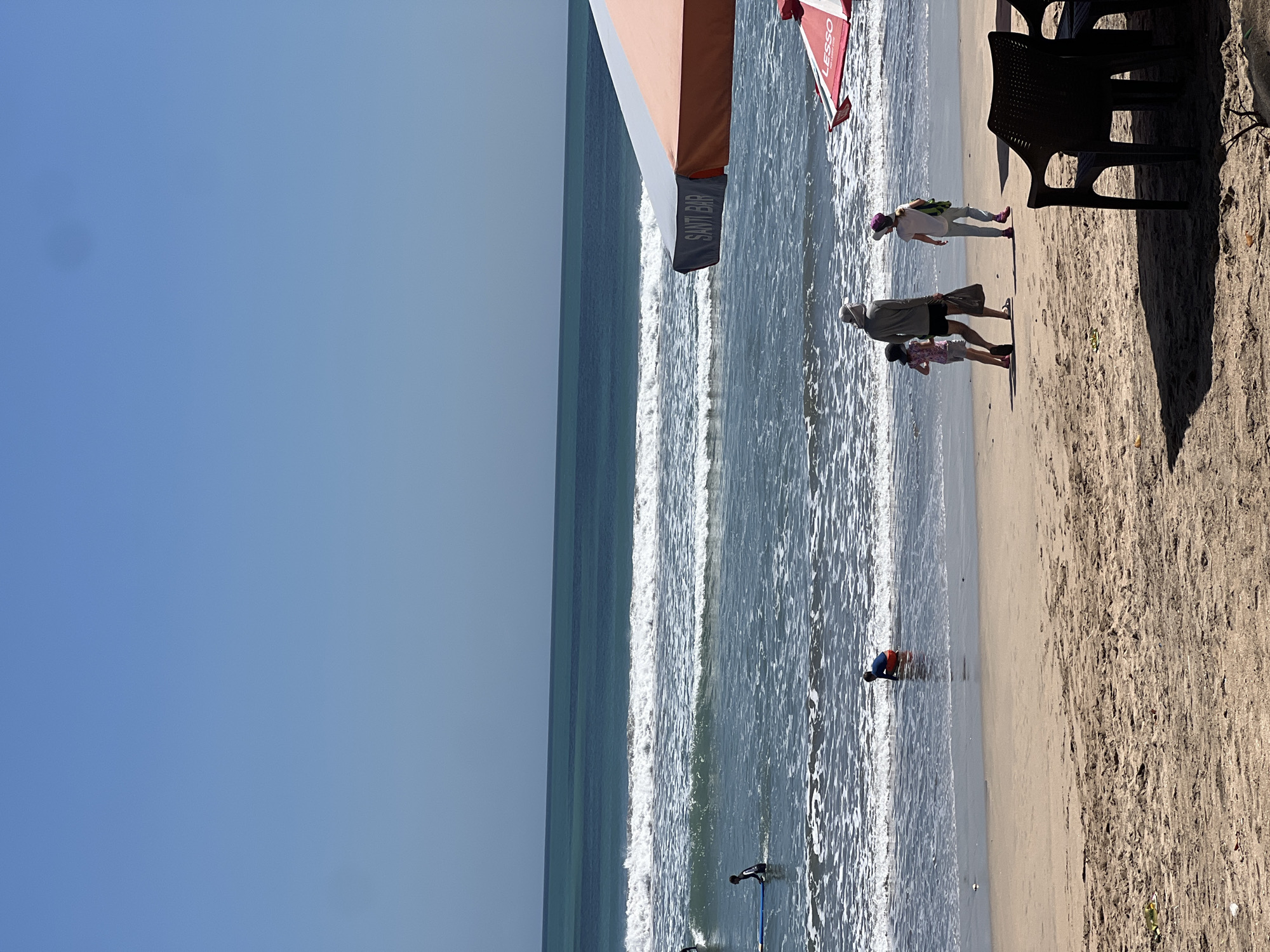This image captures a sunny day at Kuta Beach, Indonesia. The scene unfolds with a clear blue sky meeting a turquoise ocean with moderate waves. Several people are visible enjoying the beach. In the shallow water near the shore, a person in a blue shirt and red shorts is kneeling. Further out, a person in a black wetsuit is sitting on a surfboard. On the sandy beach, a group of three people are walking away from the viewer. The group consists of two children and an adult wearing a hat. To the right, two beach umbrellas are partially visible. One umbrella is predominantly orange and white, with the text "SANTI BAR" visible on its side. Beneath it, a red umbrella with the text "LESSO" is also present. In the foreground, two dark plastic chairs are positioned, casting shadows on the sand. The sand itself shows footprints and some debris.