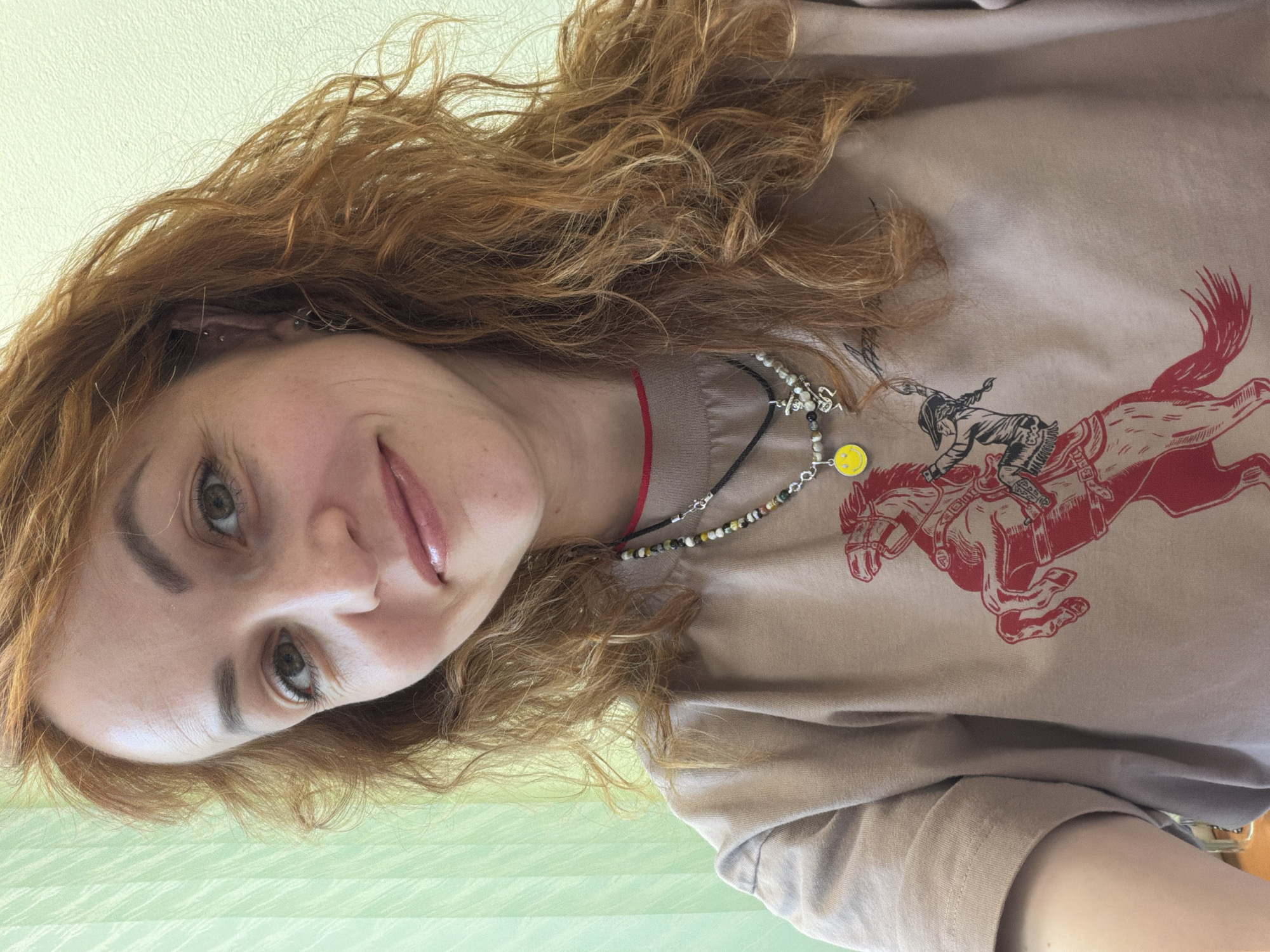 A woman with curly red hair is wearing a beige t-shirt with a red print of a rider on horseback. She is also wearing multiple necklaces, one with a yellow smiley face pendant. The background is a light green wall. The photo appears to have been taken indoors. There is no information available to determine if the image was taken in the city of Chaikovsky, Russia.