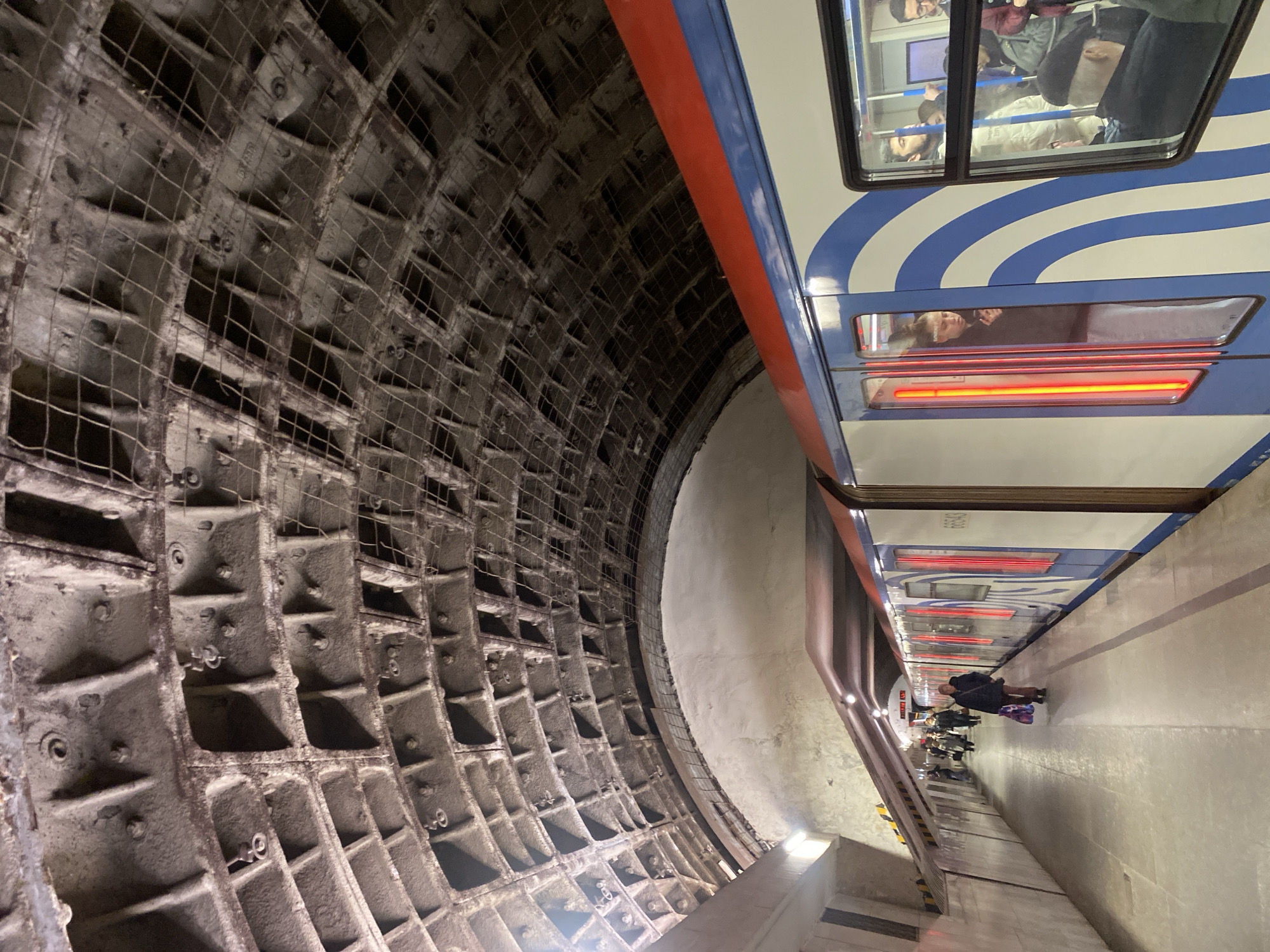 This image captures a deep underground metro station in Moscow, Russia, from a unique, tilted perspective. The camera is oriented vertically, presenting the curved tunnel as a massive wall on the left, while the train and platform occupy the right side of the frame, extending downwards.
On the left, the tunnel wall reveals a robust construction of large, interconnected gray segments, likely cast iron or concrete, forming a distinct grid-like pattern. Numerous bolts and circular rings are visible, securing these heavy components. A fine, dark mesh netting overlays parts of this structure.
To the right, a modern metro train is stopped at the platform. Its side features a distinctive livery with white, blue wavy stripes, and a prominent red horizontal stripe running above the windows. Reflections of passengers, some with visible faces and upper bodies, are seen in the train's large windows, while lower windows display bright horizontal red lights. The number "60643" is visible on the train's white side panel.
The platform below the train is wide and extends into the distance, with a few commuters visible. One person in a dark coat and boots stands closer to the foreground. The platform is illuminated by artificial lights, some casting a warm glow near the base of the train. Yellow and black striped markings are faintly visible on a structure along the platform edge. The scene depicts typical commuter activity, with people waiting for or exiting the train in an underground environment. The time of day is not discernible due to the artificial lighting.