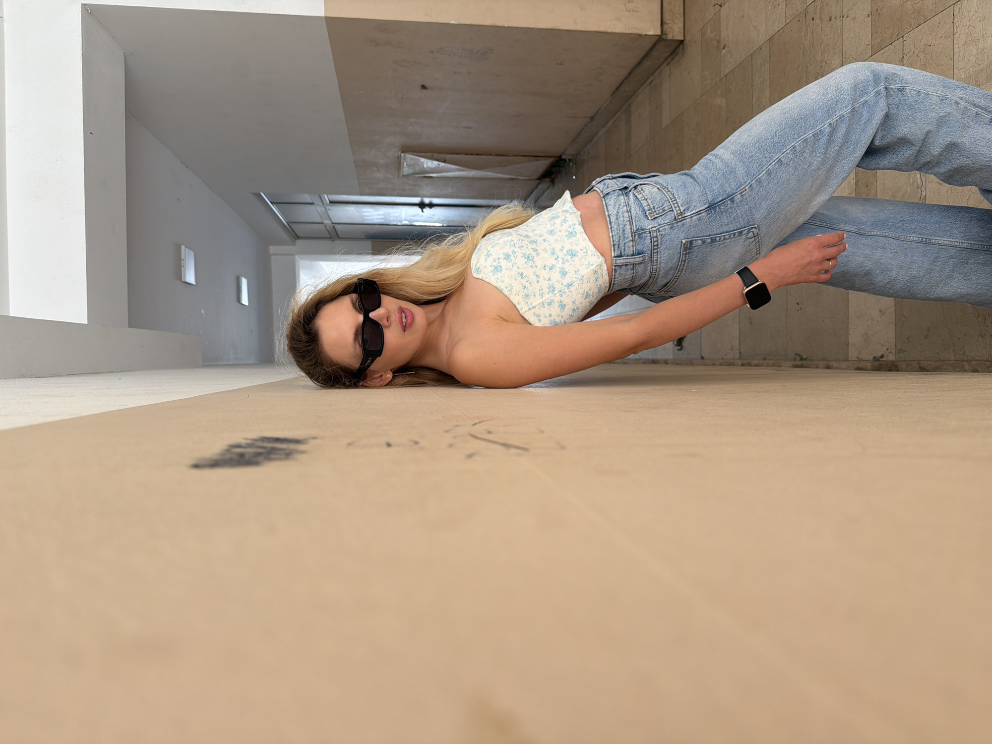 A young adult female with long blonde hair and black rectangular sunglasses, featuring a "VOGUE" logo on the left temple, poses in an indoor hallway located in Budva, Montenegro. She wears a white and light blue floral strapless crop top, light-wash wide-leg denim jeans, a black smartwatch with a gold-toned frame on her left wrist, and small gold cross-shaped stud earrings. She is leaning her right side against a faded, light-beige wall that shows faint black markings and scratches. Her head is tilted back slightly, and she looks directly towards the viewer with a neutral expression. Her left hand rests on her hip. The hallway features a light-colored, possibly beige or grey, tiled floor with visible grout lines and some discoloration. The white ceiling has multiple parallel beams and recessed rectangular light fixtures that appear to be off. In the background, the hallway extends further, revealing a glass-paneled door with a metal frame on the left, and a partially obscured object wrapped in clear plastic leaning against a far wall on the right. The environment appears dated and utilitarian, with visible wear on the surfaces.