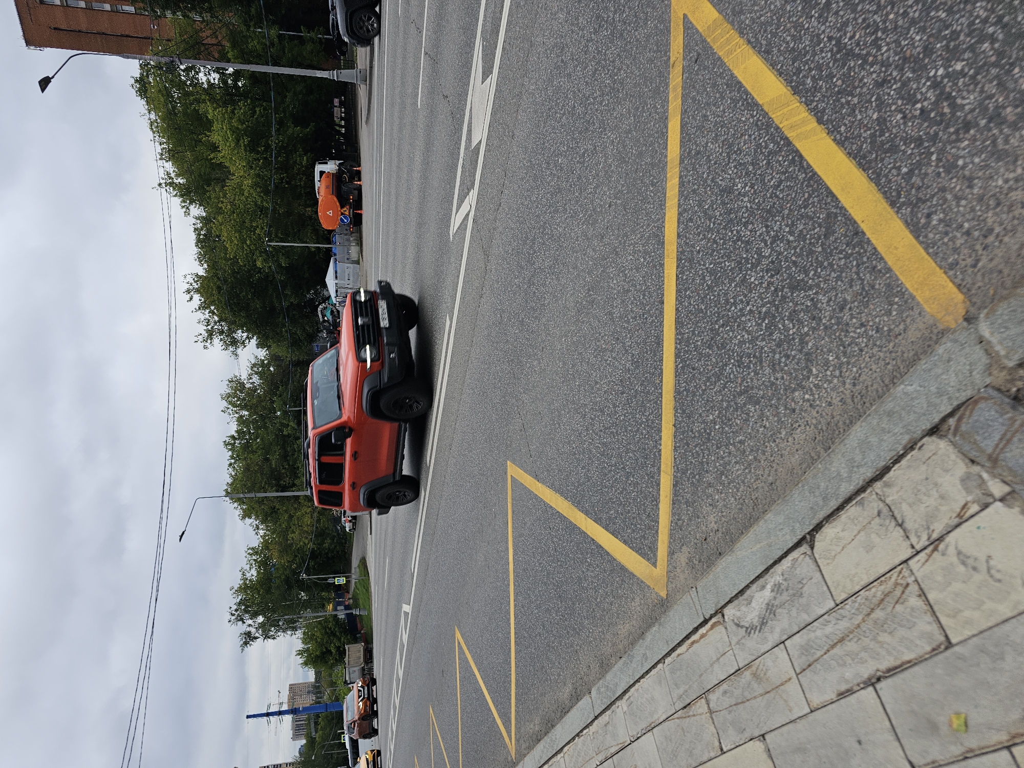 An orange off-road vehicle drives on a multi-lane asphalt road in Moscow, Russia. The road is marked with yellow lines creating a zig-zag pattern near the curb. On the left side of the road, a pedestrian sidewalk made of grey stone slabs is visible. Beyond the road, there are green trees, buildings, a street lamp, and utility poles with wires. In the background, a water truck with an orange tank and a white cab is parked on the side of the road. The sky is overcast.