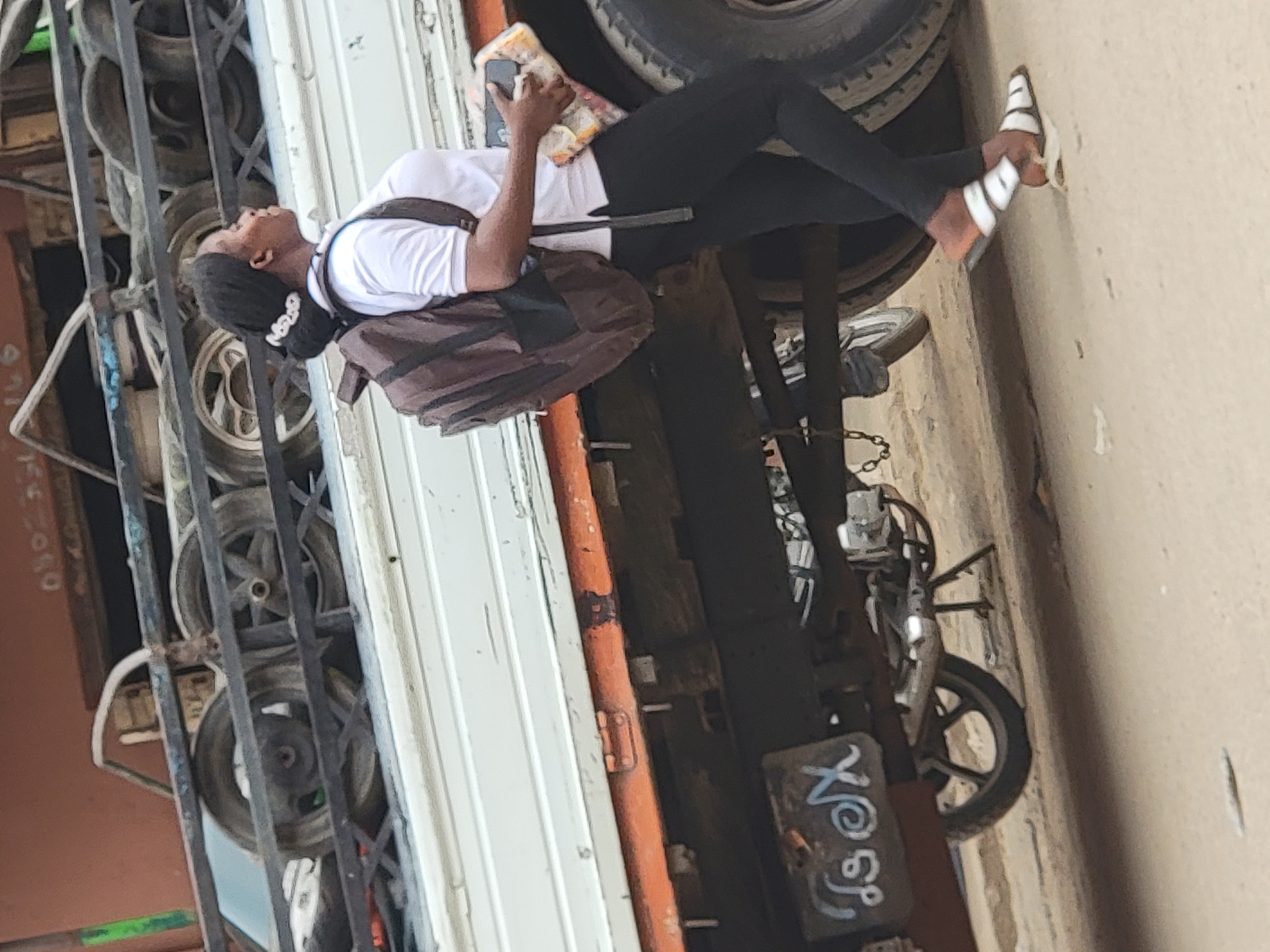 In Maiburiji, Nigeria, a young woman is depicted standing or leaning against a large white and orange truck or delivery van under bright daylight. She has dark hair tied back with a white scrunchie and is dressed in a white short-sleeved t-shirt, black leggings, and distinctive white and black striped sandals. A dark brown backpack is slung over her shoulders. She holds a smartphone in her left hand and a packaged item, possibly a snack or food, in her right. Her head is tilted back, and she gazes upwards with a neutral expression.
To the right of the woman, a motorcycle is parked, displaying a license plate with graffiti-style text that reads "X@osY". On the left side of the image, behind a dark metal gate, several vehicle tires are stacked, suggesting a tire shop or storage area. Faint, reddish numbers "040" are visible on a surface behind the gate. The scene takes place outdoors, hinting at an urban or industrial environment, possibly a street or commercial lot.