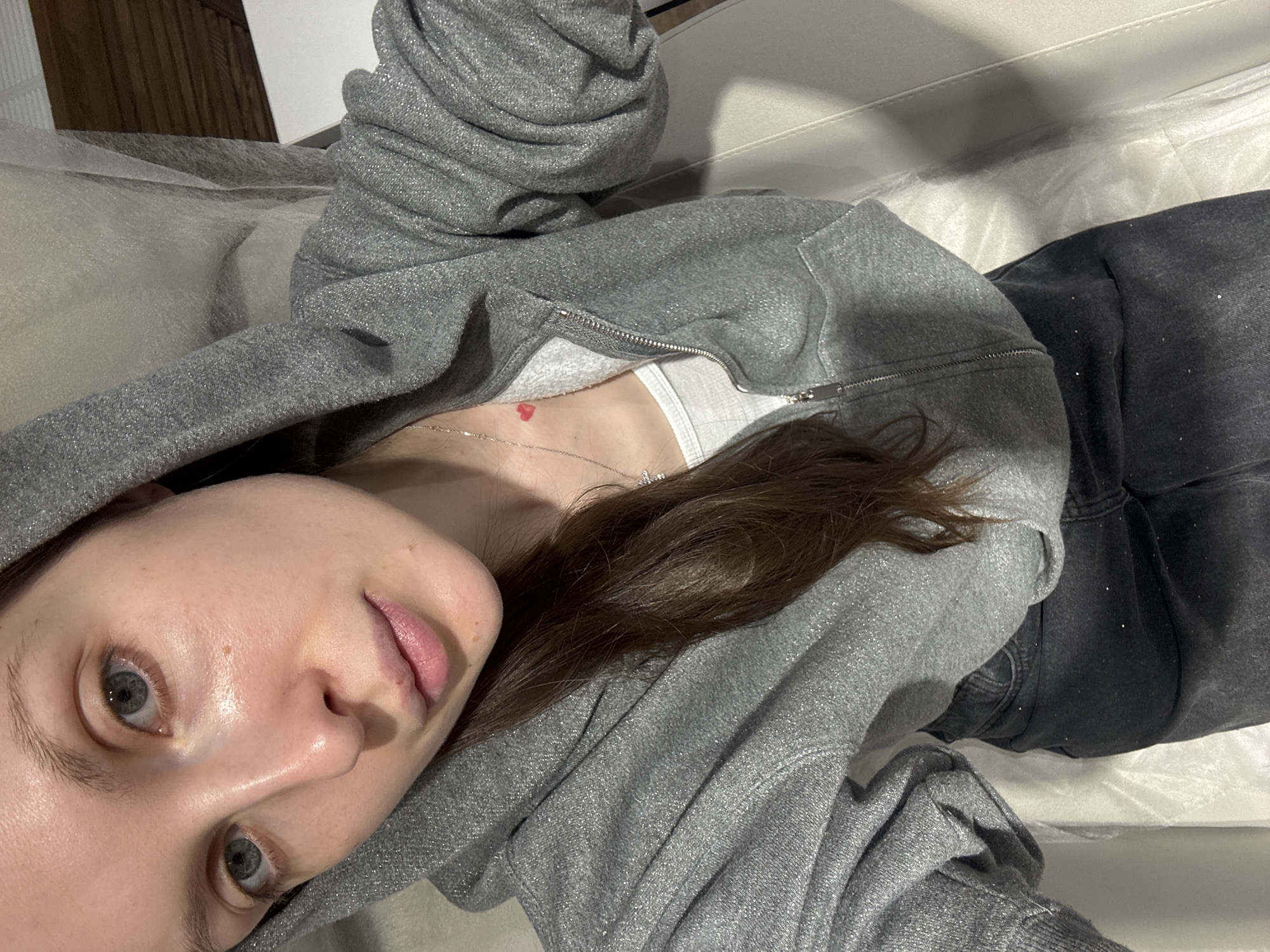 A young woman is captured in a close-up, eye-level shot, appearing to be lying down indoors in what might be Moscow, Russia. She is looking directly at the camera with a neutral and relaxed expression. Her skin is fair, and her eyes are a striking blue-grey. Her dark brown hair is visible on her right side, resting on her shoulder.
She is dressed in a casual yet stylish outfit. She wears a grey hooded sweatshirt or jacket that is partially unzipped, revealing a white top underneath. Both the grey hoodie and her dark grey or black pants appear to have a subtle shimmer or sparkle woven into the fabric. Around her neck, a delicate silver chain holds a small, distinctive red heart pendant, which is clearly visible just above her collarbone.
The immediate setting suggests an indoor environment, possibly a bedroom or living room, with a light-colored surface (perhaps a bed or sofa covered in light upholstery or bedding) beneath her. In the background, a darker, possibly wooden or paneled surface can be seen. The lighting appears to be artificial, casting soft shadows, and the overall ambiance is calm and personal. There is no visible text in the image.