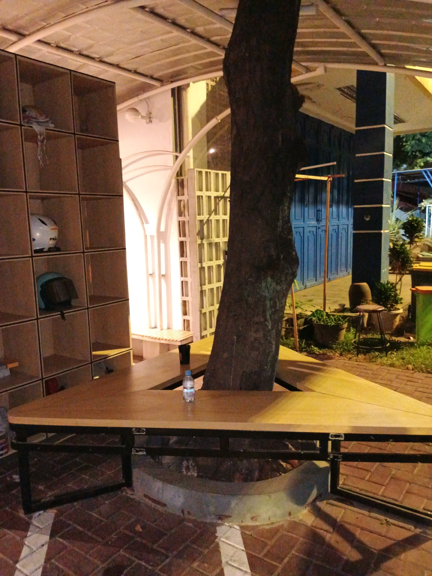 An outdoor seating area features a triangular wooden table with a metal frame, positioned around a large tree trunk. The table has a bottle of water on its surface. To the left, a tall, multi-compartment wooden shelf unit is visible, containing two helmets. In the background, a blue double door is set within a building with dark blue columns. The ground is paved with brick. The scene suggests a cafe or public space in Bandung City, Indonesia, possibly at dusk or night due to the lighting.