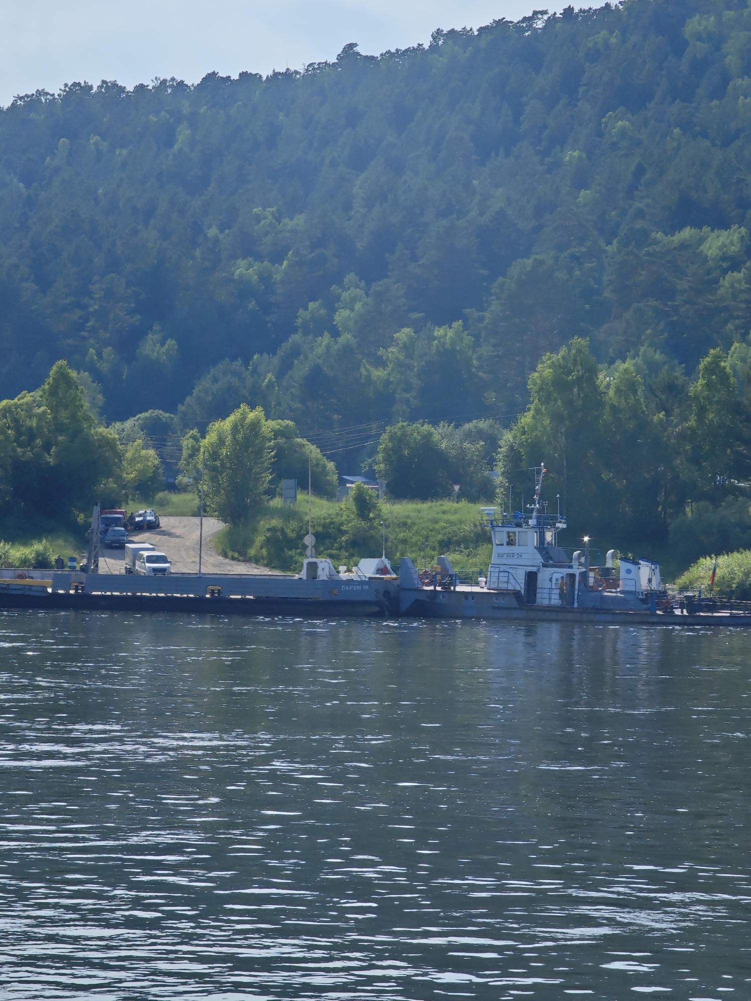 This image captures a ferryboat docked at a riverbank in Pristan', Russia. The ferry is a flat-bottomed vessel with a superstructure that houses the cabin and controls. It appears to be carrying vehicles, including a white van, a dark sedan, and a red truck, which are parked on the ferry. The ferry is positioned alongside a dirt ramp that leads up to a wooded area. A dense forest of pine trees covers a hillside in the background, with a clear sky overhead. The river in the foreground has a calm surface with ripples, reflecting the trees and sky. Two individuals can be seen on the deck of the ferry, one near the cabin and another closer to the stern. A sign on the ferry reads "ПАРОМ 94". The overall scene suggests a functional ferry service operating in a natural, forested environment.