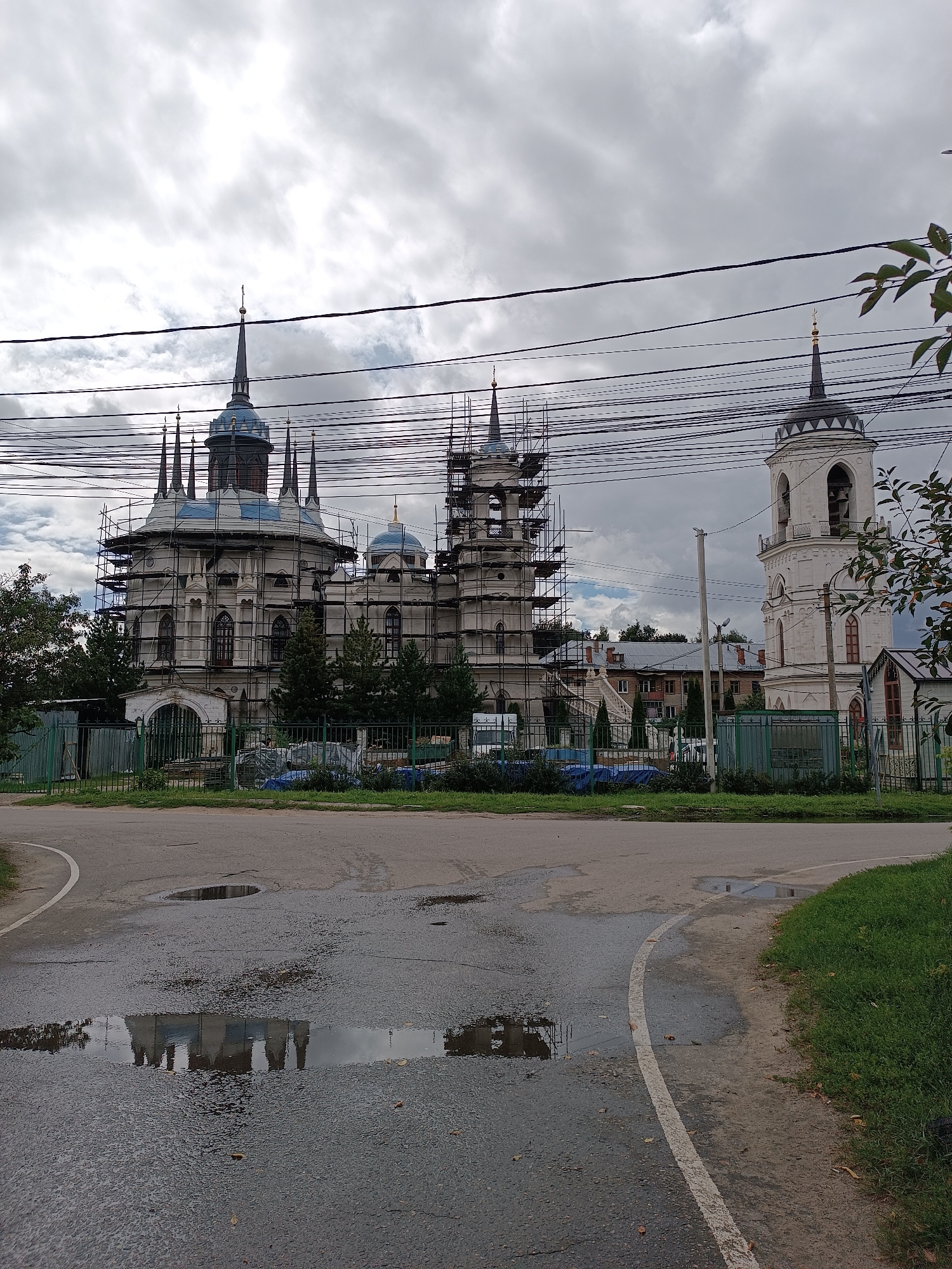 The image shows the Church of Vladimir Icon of the Mother of God in Bykovo, Russia, under renovation. Scaffolding surrounds the main church building, which features multiple spires and blue-domed roofs. To the right, a white bell tower with a dark spire stands separately. The foreground depicts a wet asphalt road with several large puddles reflecting the church structure and the overcast sky. Green metal fences line the property of the church, and some trees are visible. The overall impression is of a cloudy day with recent rain.