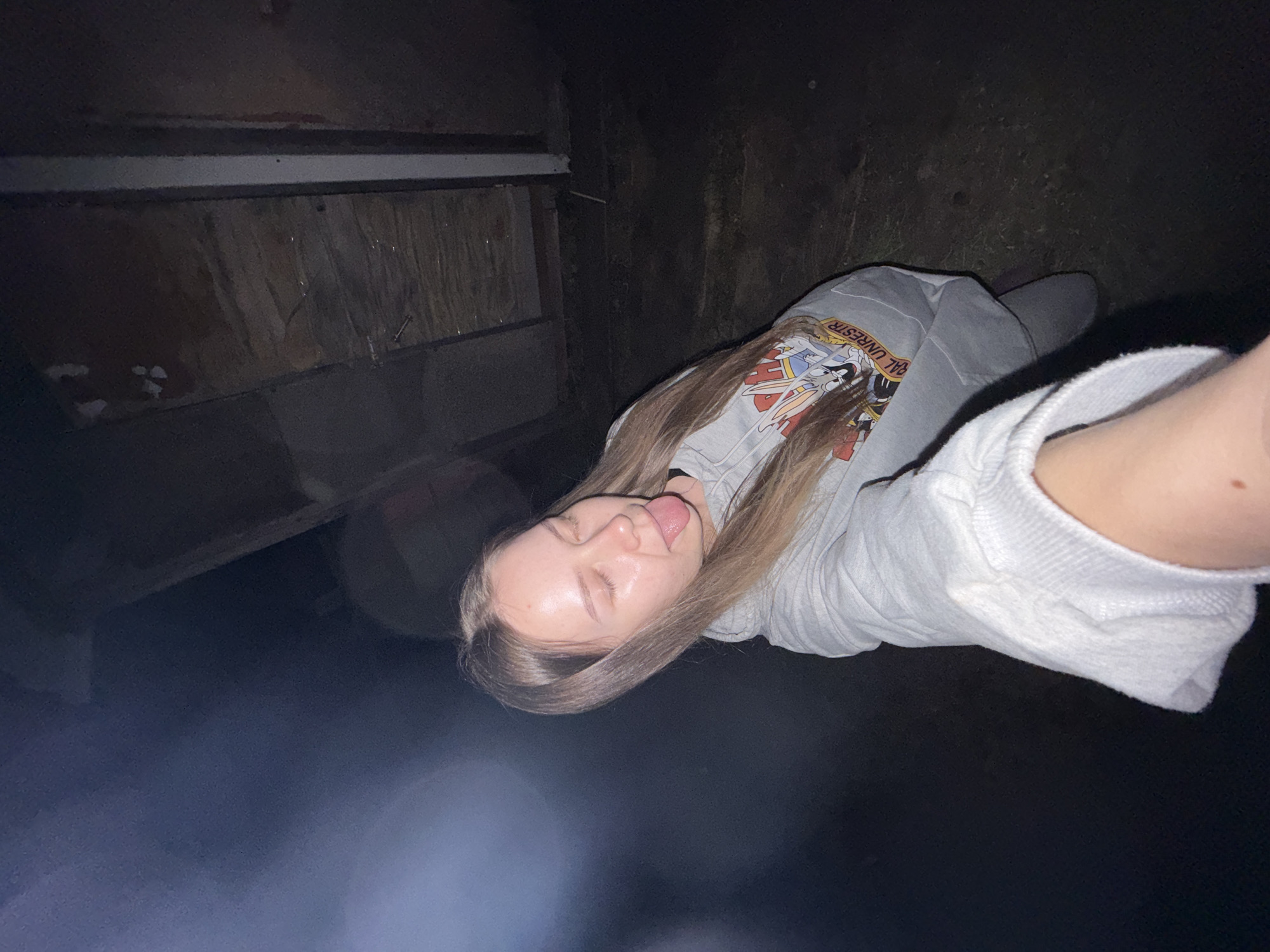 A young woman is making a selfie in a dark environment. The location appears to be Zhigalki, Russia. She is wearing a grey sweatshirt, and her hair is light brown. Her tongue is sticking out. Behind her are dark walls, possibly inside a shed or a poorly lit structure. There is some sort of shelf or storage unit to her left. The light source is unclear, but it seems to be artificial, causing glare in the bottom left corner of the image. The ground is dark and hard to discern.