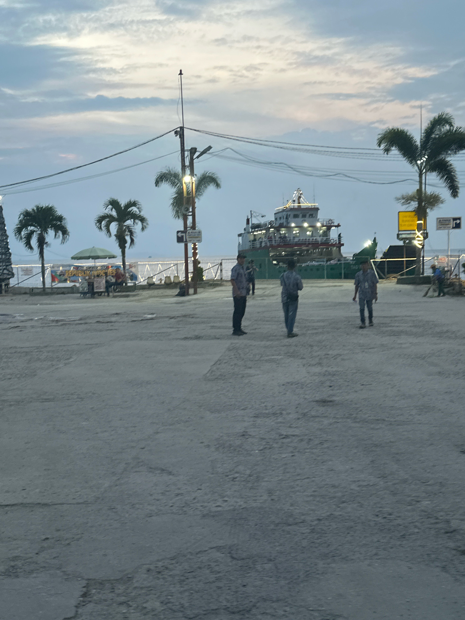 In Parsaoran Ajibata, Indonesia, an outdoor scene unfolds under a twilight sky with scattered clouds. A large, green and white ferry is docked in the background, its lights illuminated. In the foreground, a dusty, open area is occupied by several individuals. Three men in casual clothing stand in the mid-ground, appearing to be conversing. Further back, a few people are visible near a beachside area with umbrellas and stalls. Palm trees are scattered throughout the scene, framing the view of the water and the ferry. The overall impression is a bustling harbor area as day transitions to night.