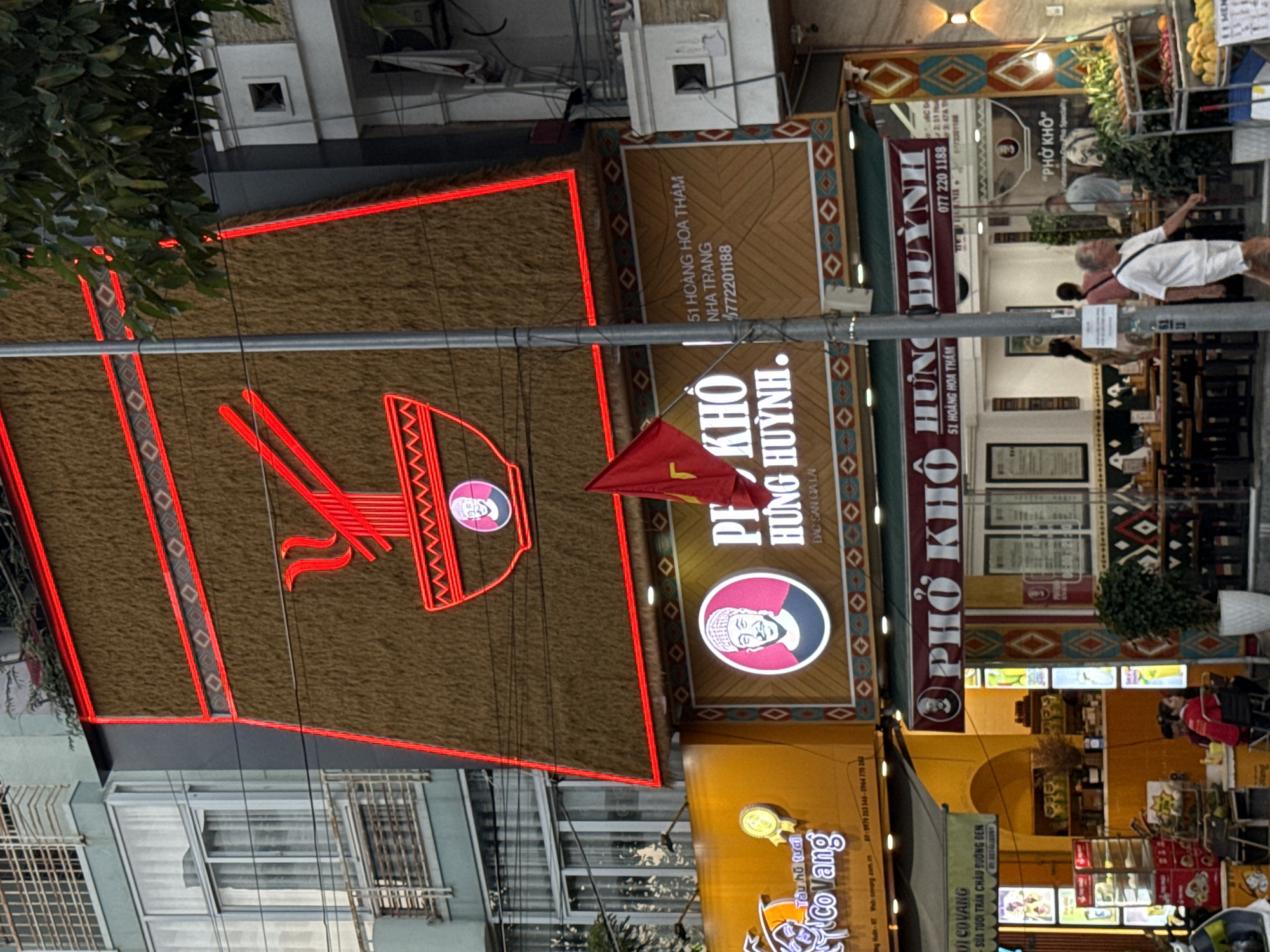 This image captures the exterior of a restaurant in Nha Trang, Vietnam, featuring a prominent neon sign and traditional Vietnamese architectural elements. The restaurant's facade is adorned with a large, stylized red graphic of chopsticks and a bowl, outlined in vibrant red neon. The main sign reads "PHỞ KHÔ HÙNG HUỲNH." Below this, smaller text indicates the address as "51 HOÀNG HOA THÁM, NHA TRANG" and provides a phone number, "0772201188." The restaurant's interior is visible through large windows, showing tables and chairs set up for dining, with patrons seated inside and a few individuals walking past on the street. A Vietnamese flag is also displayed prominently. To the left, another establishment with a yellow awning and "CoVang" branding is partially visible, suggesting a commercial streetscape. The overall scene depicts a bustling urban environment characteristic of Vietnam's coastal cities.
