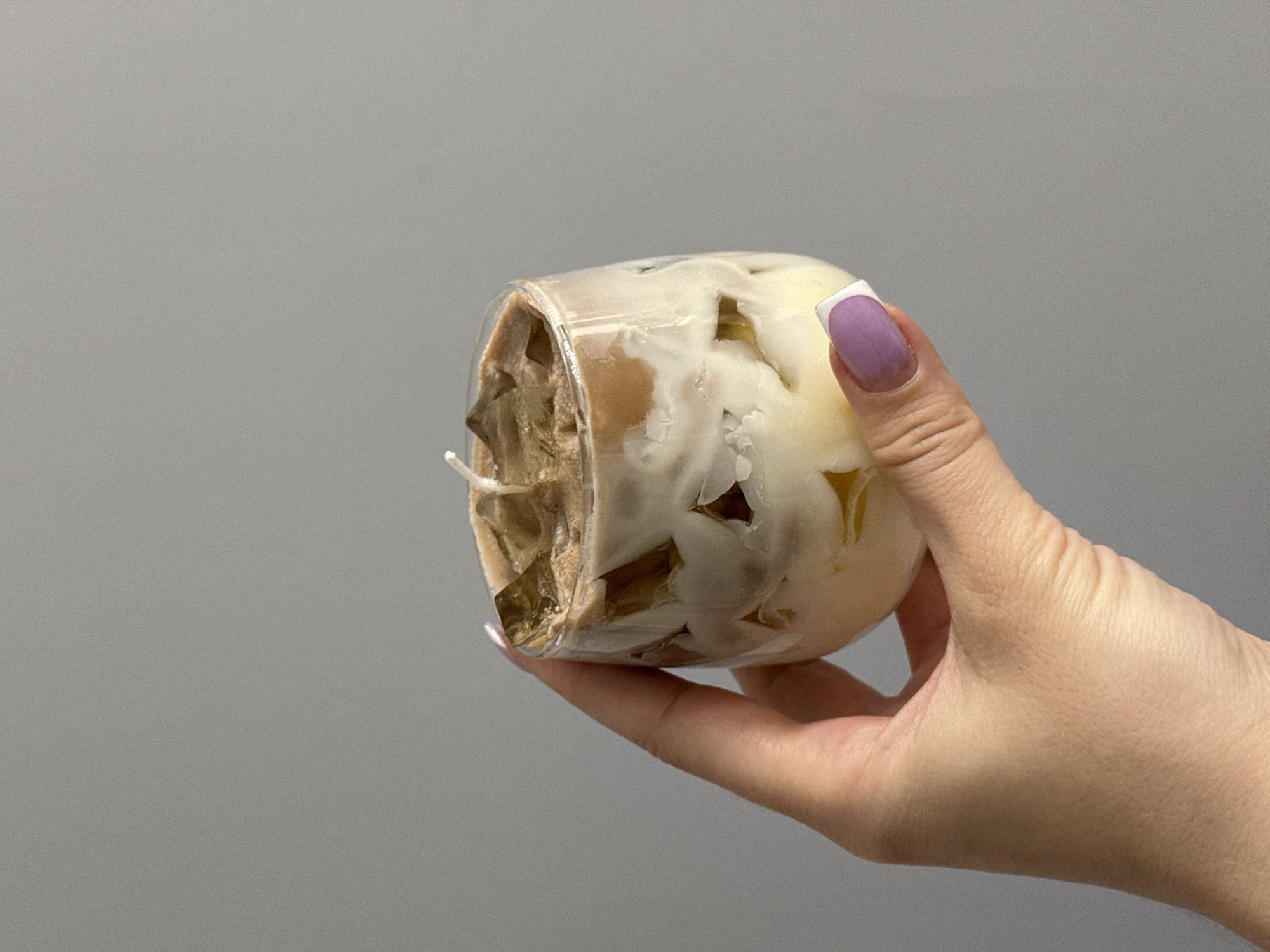 A hand with purple and white tipped fingernails holds a clear glass containing a layered candle designed to resemble an iced coffee drink. The candle has a white base layer, interspersed with brown chunks, topped with a creamy white layer, and finished with brown, ice-cube-like shapes and a light brown layer. A white wick protrudes from the top center of the candle. The background is a solid, neutral gray wall. There is no discernible location in Łódź, Poland.