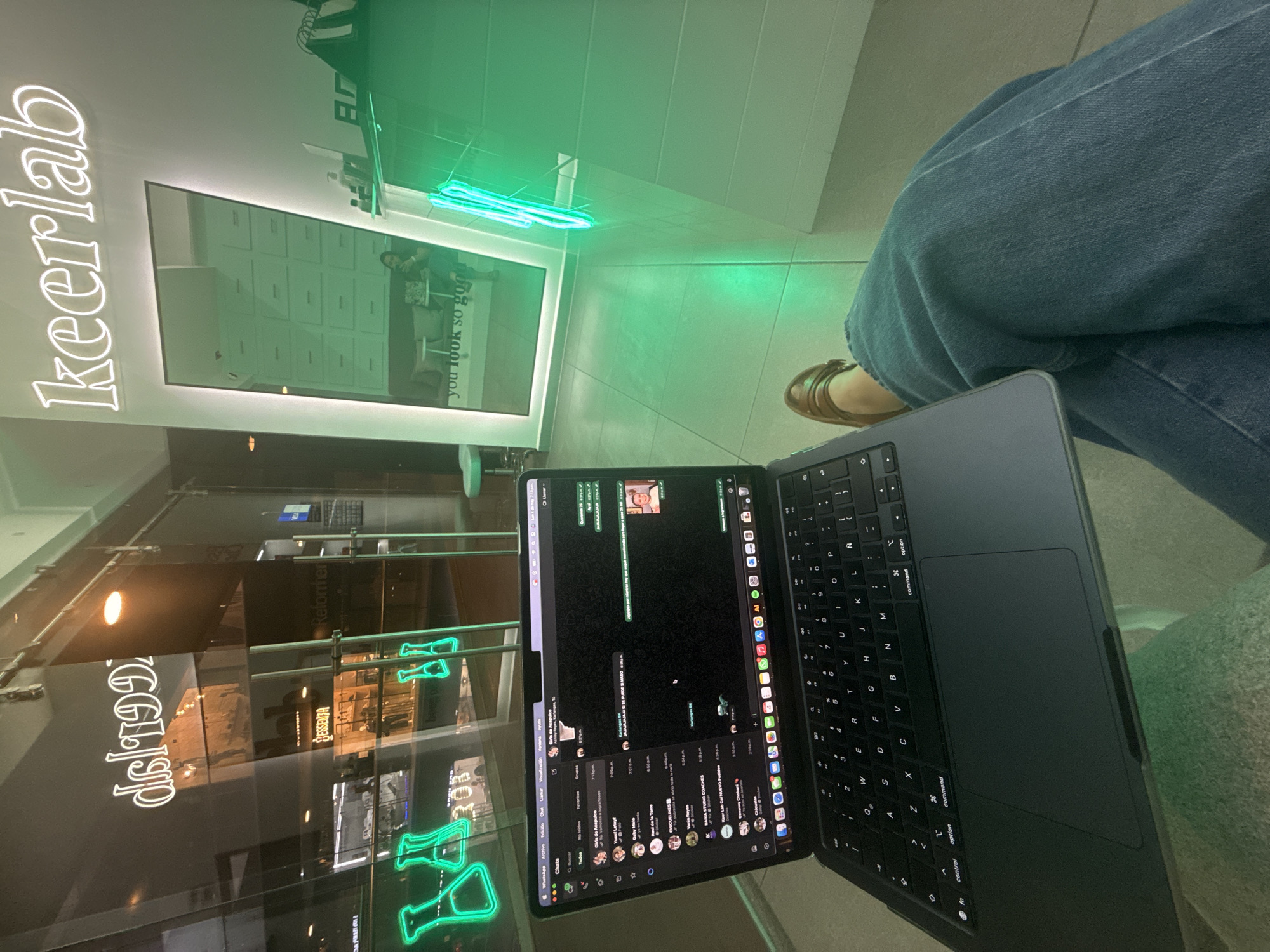 The image captures a scene inside a Keerlab location, likely in San Bernardino Tlaxcalancingo, Mexico. Visible through the window is a reflection of the store "dsl192" and "Reformher" a clothing boutique. The focus is on a person seated with a laptop open, displaying a WhatsApp chat window. The person is wearing jeans and sandals. A neon sign with the word "Keerlab" is visible near a large display screen showing the words "you look so good". There's also a green neon light fixture overhead.