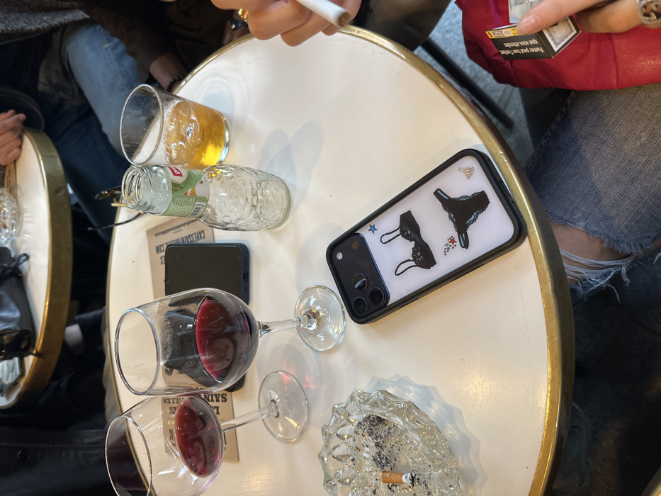 The image displays a detailed, close-up view of a round, white table with a gold-colored rim at a cafe or bar in Paris, France. The location is confirmed by a visible menu card partially reading "CAVES SAINT GILLES" and "LES HALLES," and a red cigarette pack with French health warnings.
On the table, there are two wine glasses, each containing a small quantity of red wine. A clear glass ashtray holds an extinguished cigarette butt and ash. An empty, clear glass bottle with a green label, indicating "GRENADINE," stands next to a clear glass filled with a golden beverage and ice. Two smartphones are present; one is black and placed face-down, while the other, an iPhone model, rests face-up in a clear case decorated with black lingerie motifs (a bra and panties) and small stars.
Partial views of at least three individuals are discernible. In the foreground, the legs of a person wearing distressed blue jeans are visible, with rips at the knees. This individual's left hand, adorned with a ring, is seen holding a white cigarette and the red cigarette pack. Another person's lower torso and legs, also dressed in blue jeans, are visible seated on the far side of the table. A third person's arm is faintly visible in the upper right background.