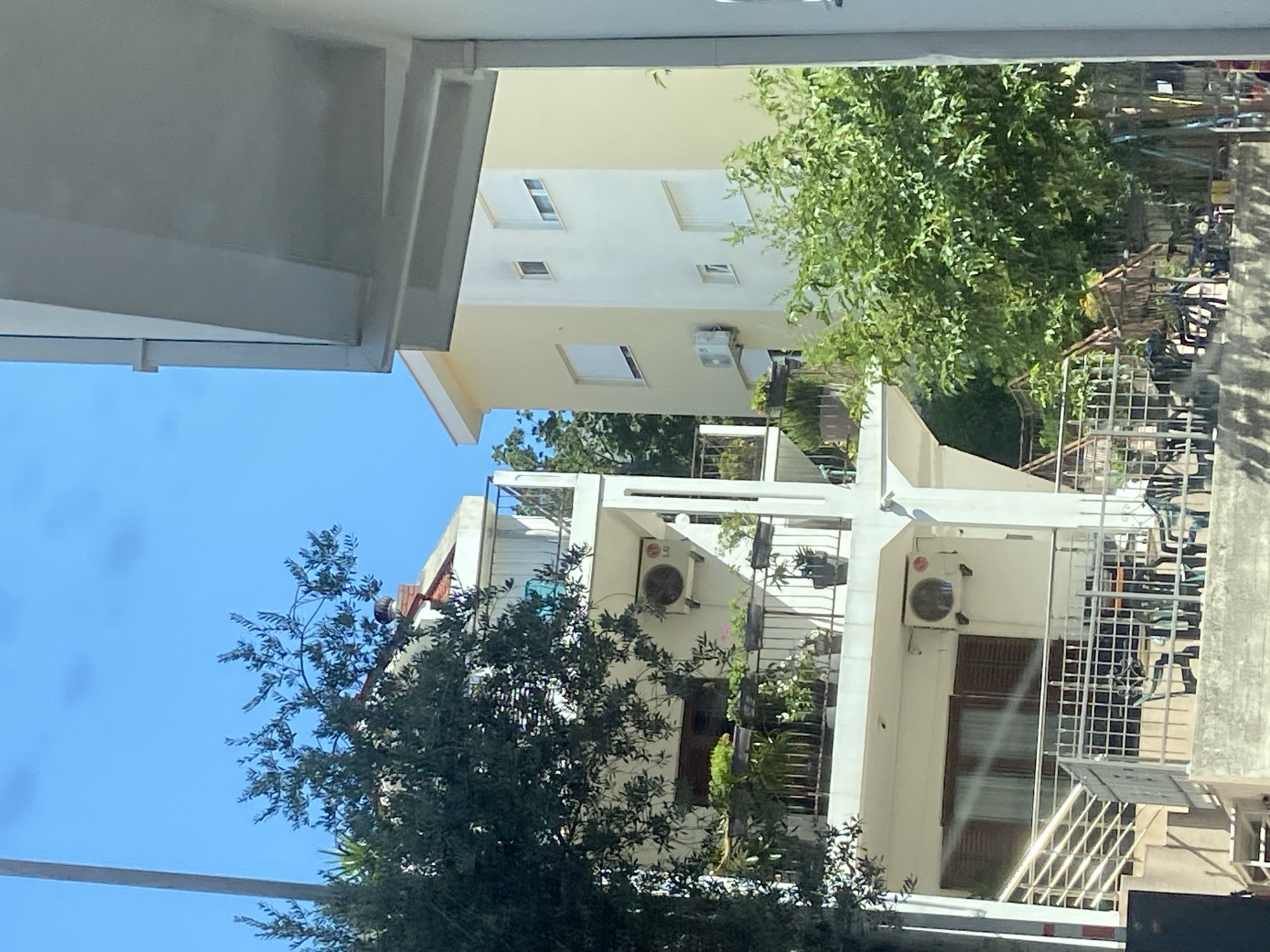 This is a daylight, medium-distance, eye-level, exterior, full shot of a residential building in Budva, Montenegro. The building is white with a balcony and planters, and a yellow building is visible behind it. There are trees and a clear blue sky. There are no people visible. The scene is tranquil.