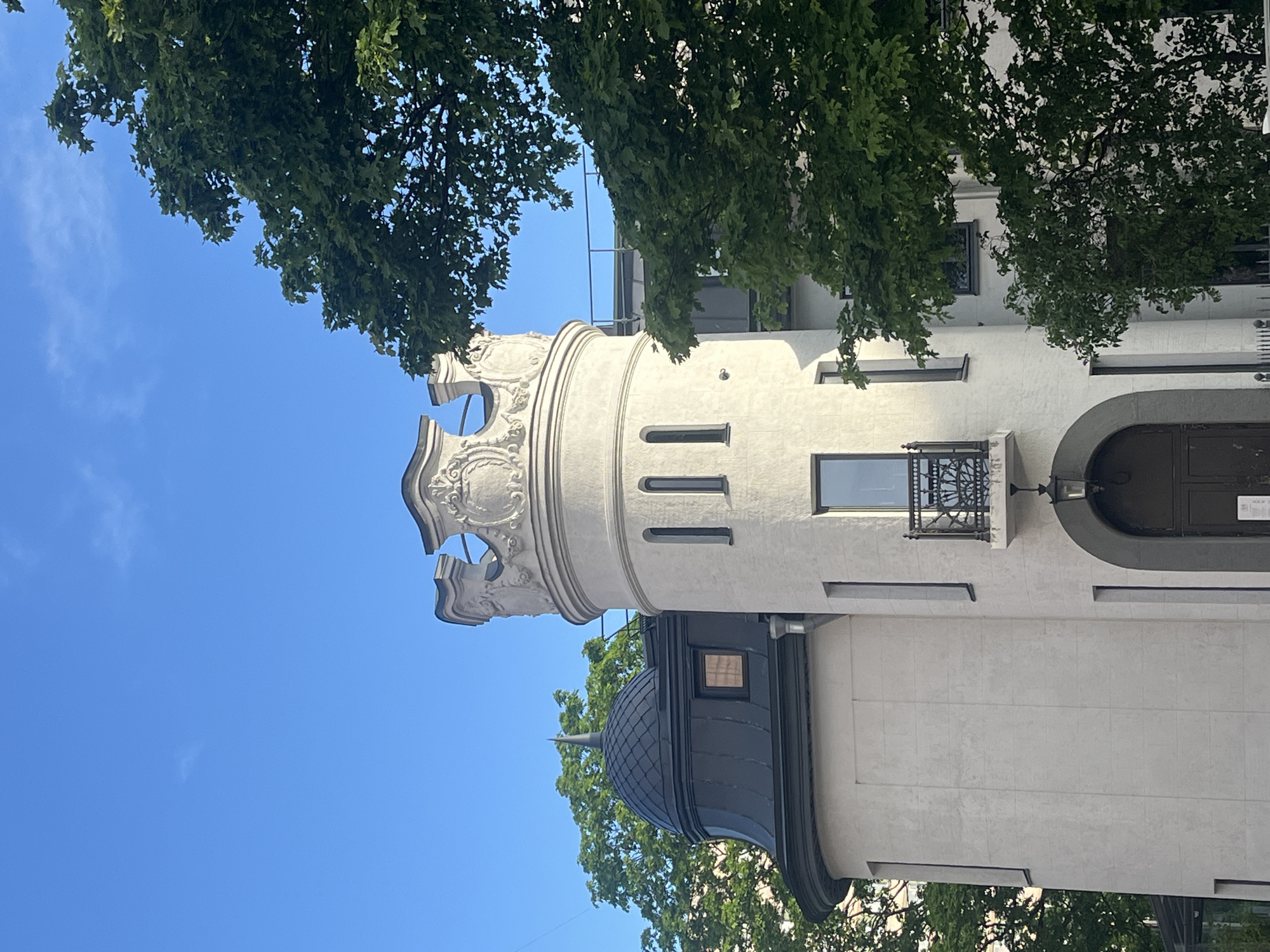 The image displays a detailed architectural view of a white building, likely a historic edifice in Saint Petersburg, Russia, under a clear blue sky. The central feature is a prominent cylindrical tower section, crowned with an ornate, carved frieze and decorative elements that suggest a stylized crenellation. Several narrow, vertical windows are visible on the tower's facade. To the right of the tower, a flat wall section features a larger rectangular window adorned with a small, dark, wrought-iron balcony. Further right, a dark, arched doorway can be seen. Below the main tower, a dark, dome-shaped roof with a pointed finial is partially visible. The building is partially framed by vibrant green tree foliage, especially at the top and left, contrasting with the bright white stone. The sky is a vivid blue with a few faint wisps of clouds, indicating clear, sunny weather during daylight hours. No people or specific activities are depicted. No discernible text is visible within the image.