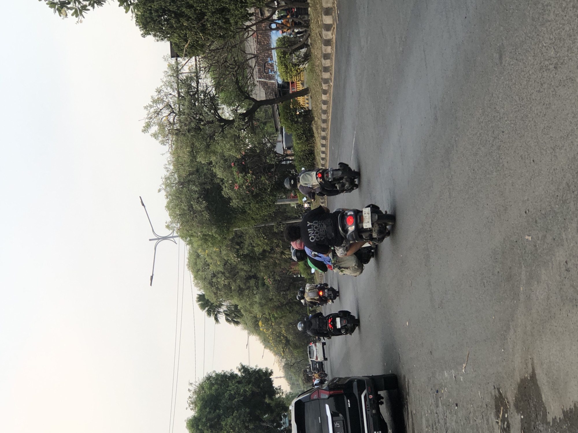 The image captures a street scene in Ngaban, Indonesia, likely during the late afternoon or early evening due to the ambient light. A black SUV is parked on the left side of the asphalt road, with its rear facing the camera. Several motorcycles are traveling on the road, mostly from right to left in the frame. One motorcyclist, in the foreground, is wearing a black t-shirt with the text "ONLY GOOD VIBES" printed on it, and is riding a scooter with a red taillight. Behind him, other motorcyclists are visible, some with passengers. Trees with green foliage line both sides of the road, and a street lamp is visible in the upper portion of the image. The sky is overcast with a light gray hue. The overall impression is one of everyday traffic in a tropical urban or suburban environment.