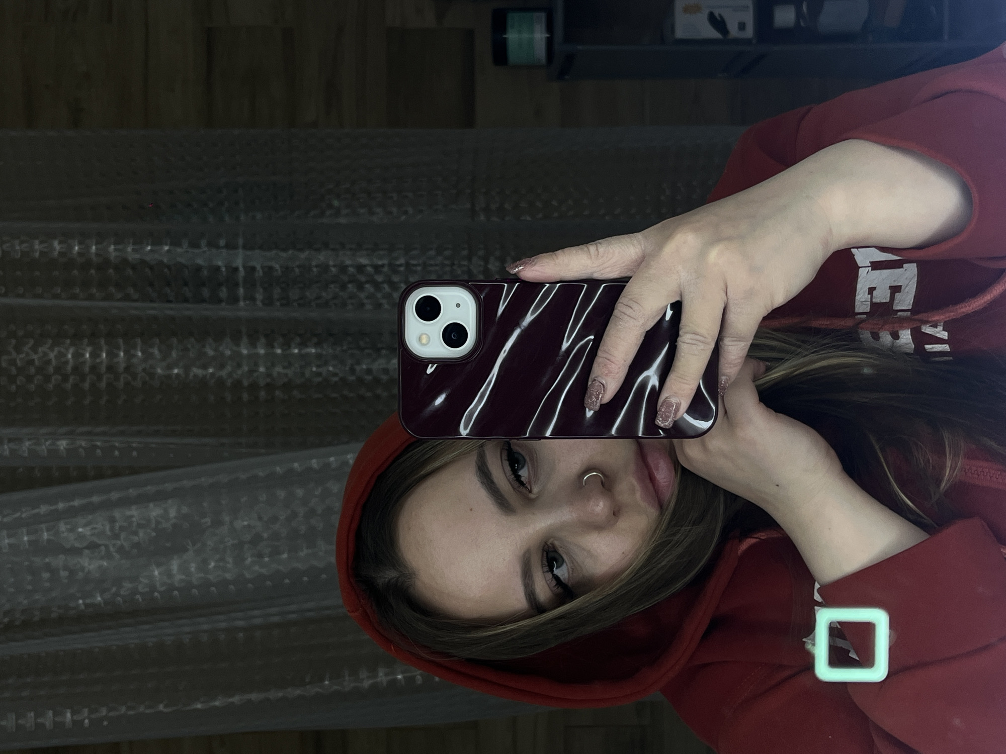 The image, taken in Krasnodar, Russia, captures a young woman from the waist up, taking a mirror selfie. She is wearing a red hooded sweatshirt, with white lettering partially visible on the front that reads "VE". Her long brown hair frames her face, and she has a nose ring on her left nostril. She looks directly at the camera with a neutral expression.
She is holding an iPhone in her left hand, which is encased in a dark red or maroon case featuring a glossy, wavy pattern. Her nails are manicured with a sparkly polish. On the cuff of her right sleeve, a distinctive glowing square-shaped accessory is visible.
The background behind her consists of a highly textured, semi-translucent material, possibly a shower curtain, privacy screen, or patterned wall. Above this, a darker area reveals shelves with dimly lit objects, including a green container and a white box. The overall lighting is dim, suggesting an indoor setting with subdued illumination. No specific time of day or weather is discernible.