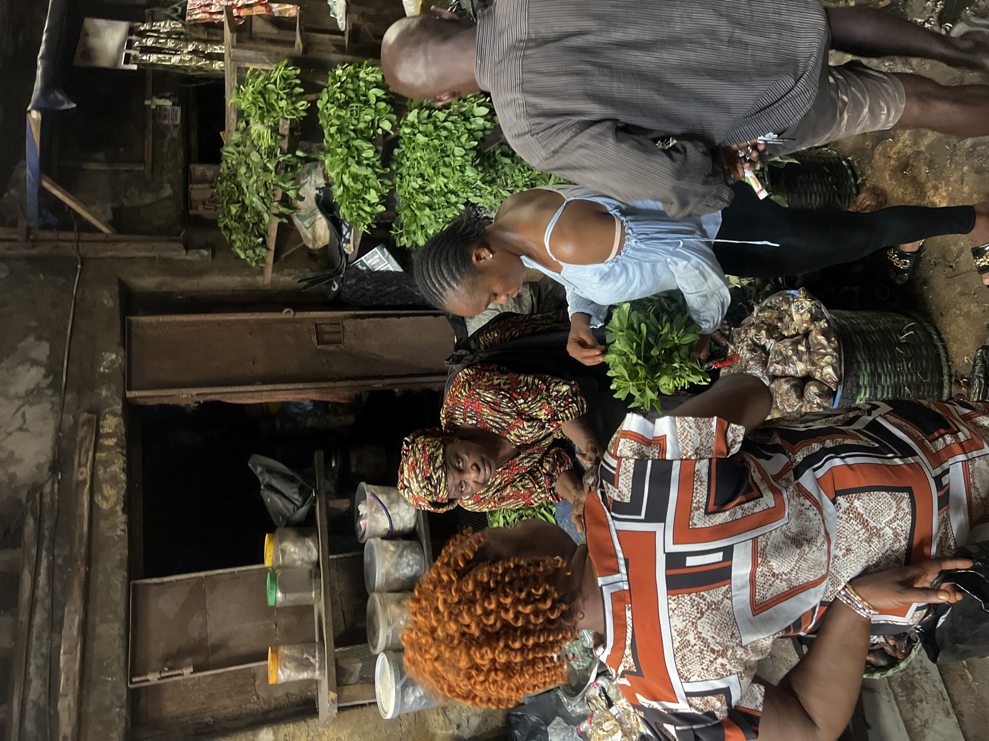 In Nkpor, Nigeria, a marketplace scene unfolds as three individuals, two women and one man, engage in commerce. The woman in the foreground, with vibrant orange, curly hair, wears an orange, white, and black patterned top and examines a bunch of leafy greens. Facing her, an older woman, draped in a colorful patterned headscarf and an equally vibrant African print top, appears to be the vendor. To the right, a woman in a light blue off-the-shoulder top and black leggings holds a bunch of greens, seemingly in discussion with a man in a grey pinstripe shirt and camouflage shorts, who is looking away from the camera.
The background reveals a stall with wooden shelves laden with produce and jars. Behind the vendor, a dark opening suggests a storage area or passageway. The overall environment is rustic, characterized by the textures of worn wood, metal, and packed earth flooring, typical of an open-air market. Several baskets and bags are visible, containing more produce and goods. The lighting is dim, suggesting an indoor or covered market setting, with some overhead illumination casting shadows.