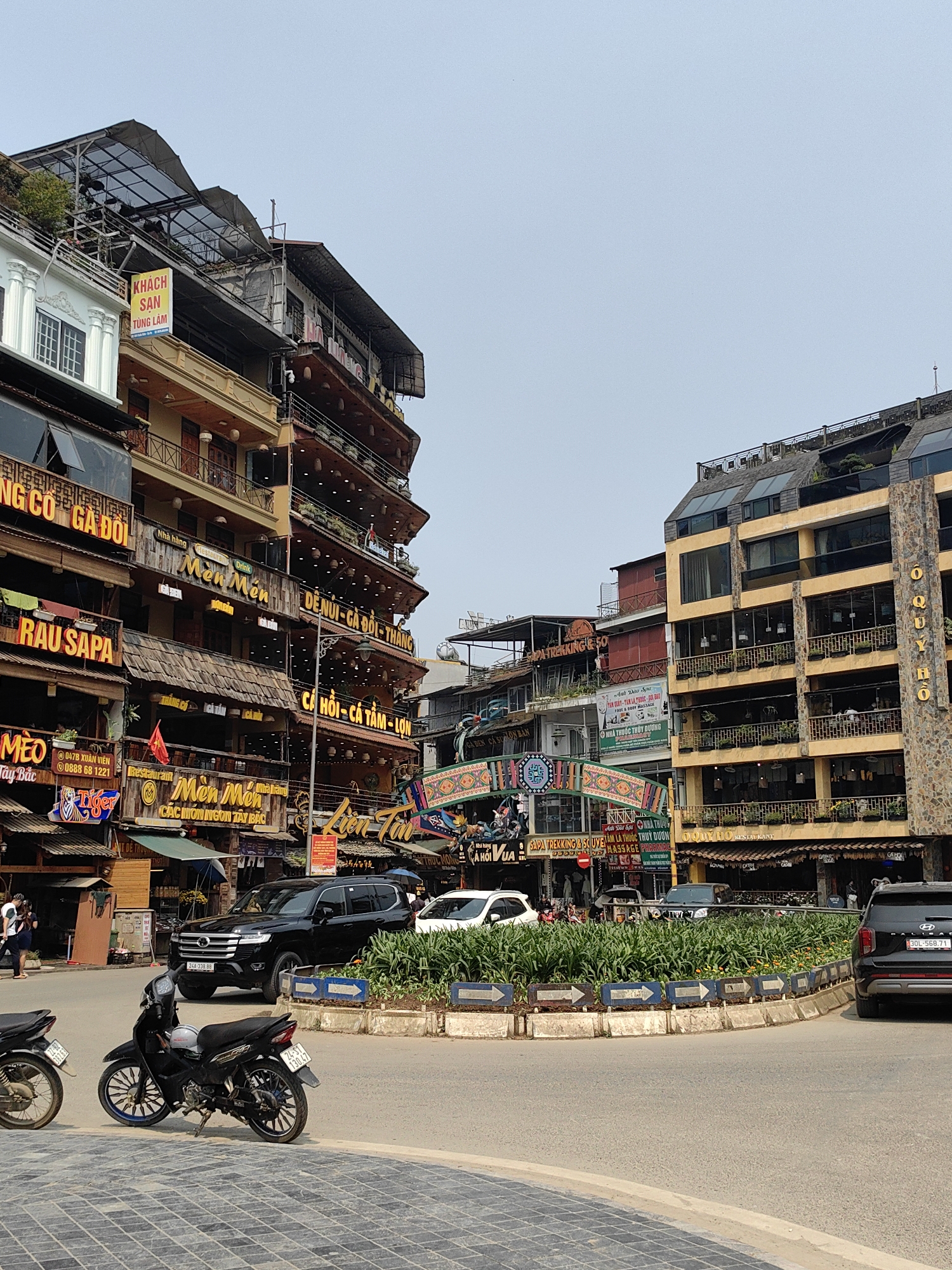 This image depicts an urban street scene in Sa Pa, Vietnam, under a clear or hazy daytime sky. The scene is dominated by multi-story commercial buildings lining both sides of a paved road.
On the left, a complex of vertically stacked buildings features numerous restaurants and a hotel. Notable signs include "KHÁCH SẠN TÙNG LÂM" (Tung Lam Hotel) at the top, and multiple "Món Mến" (Delicious Dishes) signs, along with "RAU SAPA" (Sapa Vegetables), "ĂN GÔ GÀ ĐỒI" (Eating Hill Chicken), and "CÁ HỒI - CÁ TẦM - LỢN" (Salmon - Sturgeon - Pork). These structures exhibit ornate balconies with railings, hanging lights, and some greenery. A Vietnamese flag is visible on one of these buildings.
On the right, a large, multi-story building with a light yellow facade and a prominent stone column section vertically displaying "O QUÝ HỒ RESTAURANT" features large glass windows and balconies adorned with planters.
The road features a central, circular planted island with green shrubs and small flowers, bordered by blue and white directional arrow signs. Two motorcycles are parked on the tiled sidewalk in the foreground on the left; one black motorcycle has blue accents on its wheels and a license plate 24A-330.47. A black SUV (license plate 24A-338.88) is visible on the road behind the motorcycles, followed by a white SUV. A black SUV (license plate 30L-568.71) is partially visible on the far right, facing away from the viewer.
Several individuals are present on the left sidewalk near the black SUV, including one person in a white shirt. Other indistinct figures are visible further down the street. A decorative, colorful arched structure spans across the street in the mid-ground. Utility lines crisscross overhead. Streetlights and various advertising banners are also present throughout the scene.