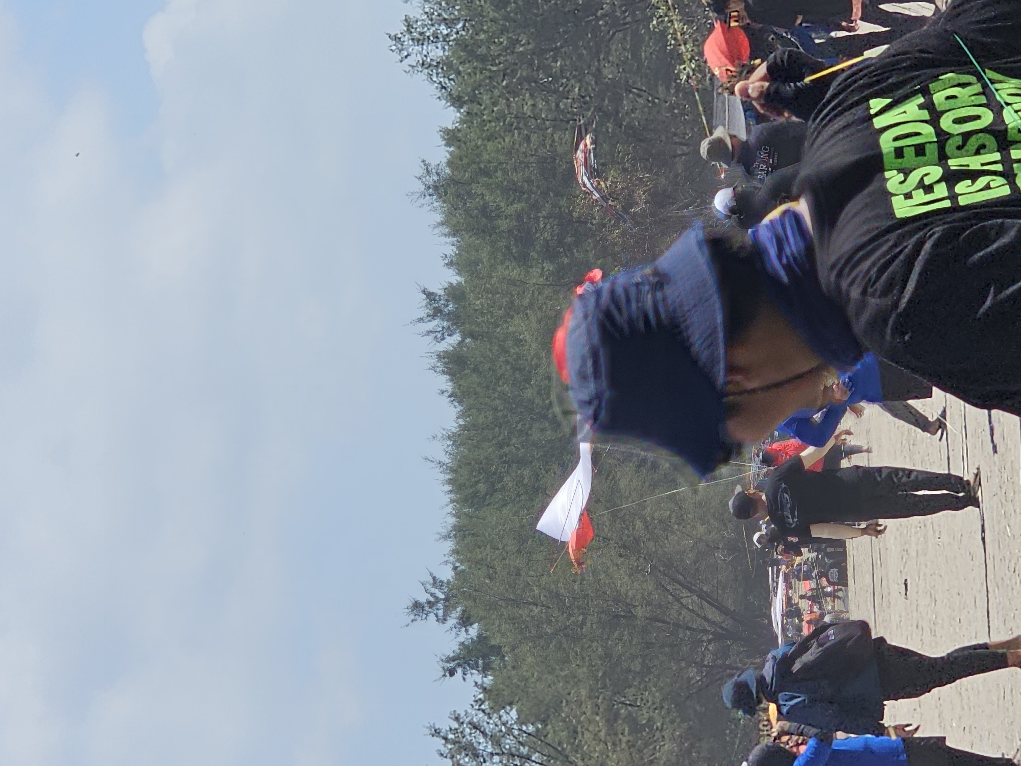 This is a medium shot of a beach event, likely a kite festival, with people, kites, and trees visible under a bright sky. In the foreground, a person wearing a dark blue bucket hat and a black t-shirt with green lettering that reads "YESEDAY IS A STORY" is partially visible from behind. To the left of this person, another individual in a blue jacket and a dark bucket hat with a backpack walks away from the camera. Further into the scene, other people are scattered across the sandy beach, some wearing hats and casual clothing. Several kites are in the air, including a prominent red and white kite with a long tail. In the background, dense green trees line the edge of the beach. The overall atmosphere suggests an outdoor gathering on a sunny day.