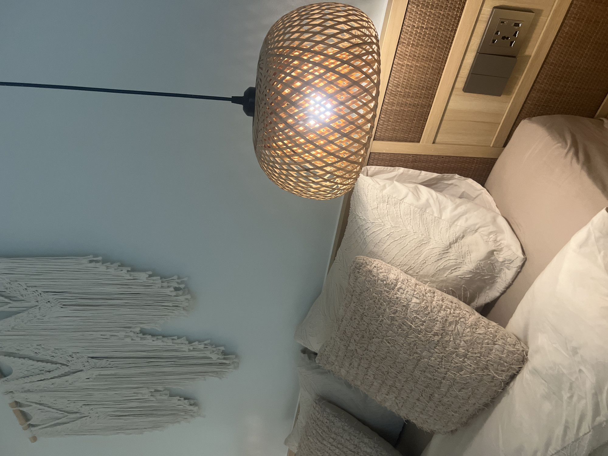 The image depicts a close-up view of a neatly made bed in a room, likely a bedroom, in Ko Samui District, Thailand. The bed is dressed with white linens and several textured throw pillows in neutral tones. Above the bed, a macrame wall hanging in white is mounted on a light blue wall. To the right of the bed, a woven pendant light with a bamboo-like texture hangs, casting a warm glow from an illuminated bulb. The headboard is made of a woven rattan material with wooden accents. On the headboard, a modern electrical outlet with multiple plug types is visible, alongside two dark-colored light switches. The lighting suggests it may be evening or a dimly lit room.