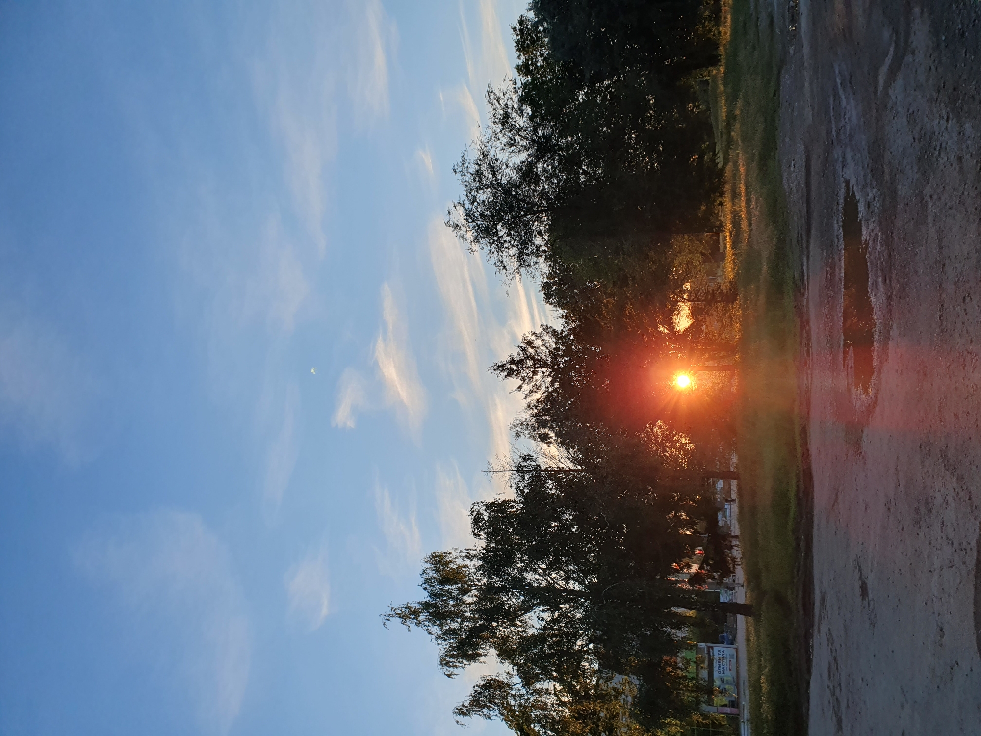The image depicts a serene sunset scene in an outdoor urban environment. The sky is a clear blue, adorned with wispy white clouds that are catching the golden hues of the setting sun. The sun itself is a bright, fiery orb, partially obscured by the dark silhouettes of a dense line of trees that stretches across the mid-ground of the image. The light from the sun beams through the foliage, creating a dramatic lens flare effect that casts a warm glow on the scene. In the foreground, a dirt or gravel path is visible, with puddles reflecting the sky and the sunset. To the left, the edge of a building with some signage is partially visible, hinting at the urban setting. The overall atmosphere is tranquil and visually striking, with a strong contrast between the dark silhouettes of the trees and the brilliant light of the sunset.