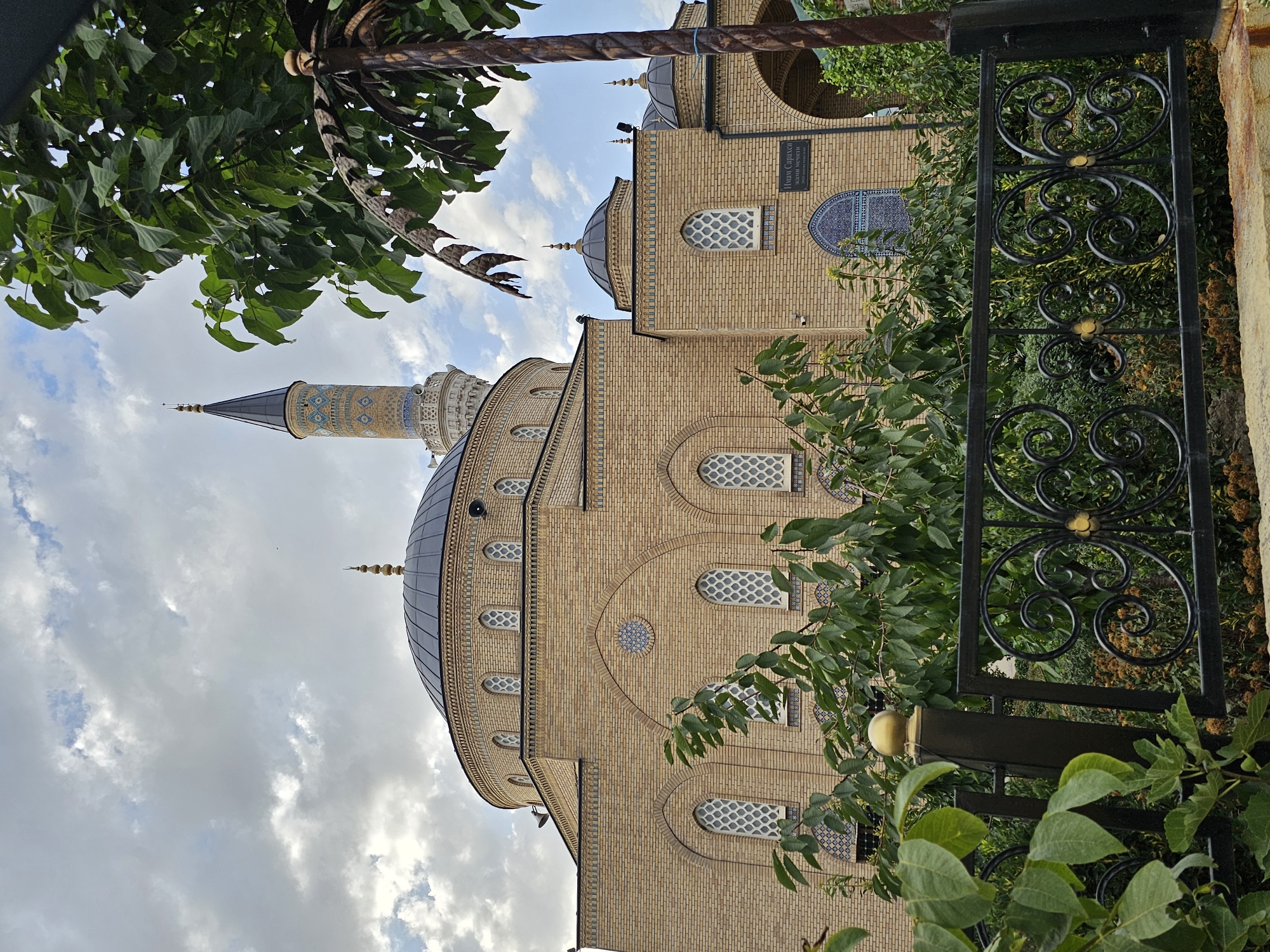 An exterior, low-angle, eye-level shot captures the Imam Sarakhs Mosque in the city of Özgön, Kyrgyzstan, under a cloudy sky. The mosque is constructed of tan brick, with a large, dark dome topping the main structure. A tall, slender minaret with ornate geometric patterns in blue and yellow tilework rises to the left of the dome. Several smaller, dark domes are visible on the right side of the building, which also features arched windows with decorative latticework. Lush green foliage partially obscures the lower portion of the mosque, with trees in the foreground and a black wrought-iron fence with gold accents running across the bottom of the frame. A sign with Cyrillic script is mounted on the mosque wall above an arched entrance. No people are visible in the image.