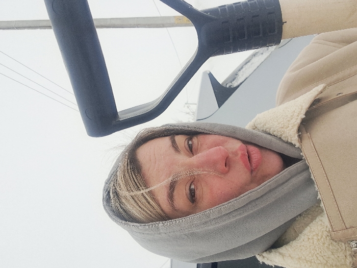 An adult female, appearing to be in her 30s-40s, is depicted in a close-up, selfie-style photograph. Her light-colored hair is partially visible beneath a grey hooded sweatshirt, with noticeable frost or ice crystals on strands near her left temple. Her face exhibits some redness, consistent with cold exposure, and her lips are slightly puckered. She is dressed in a light beige jacket featuring a cream-colored sherpa-lined collar, worn over the grey hood. In the right foreground, the black D-handle and a portion of the light-colored shaft of a shovel are visible, held by the subject. In the background, behind the subject's left side, a silver or light grey vehicle, possibly a car or SUV, is partially visible with snow accumulation on its rear window or roof. To the far right, a utility pole with overhead wires is present. The sky is uniformly white and overcast, indicating a cold, snowy, or heavily cloudy winter environment. The scene is located in the city of Полянки, Russia.