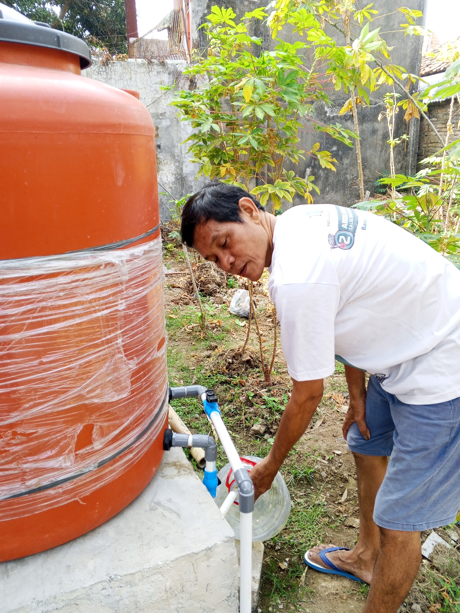 In the city of Ciruas, Indonesia, a man in a white t-shirt and blue shorts is seen tending to a large orange water tank. The tank, wrapped partially in clear plastic, sits on a concrete base. The man is bending down, holding a clear plastic container under a spigot connected to the tank via PVC pipes and fittings. The surrounding environment is outdoors, with dry soil, sparse green grass, and some leafy plants. In the background, a weathered concrete wall and a roof are visible, suggesting a residential or semi-rural setting. The man's posture and the action of filling the container indicate he is drawing water from the tank.