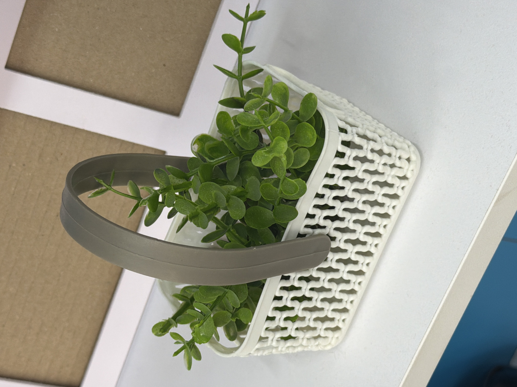 A close-up, high-angle shot shows a white woven basket filled with green artificial plants. The basket has a gray handle. In the background, two picture frames with tan inserts are visible. The basket is placed on a white surface, which appears to be a shelf or table. The lighting is bright and even, with a slight shadow cast by the basket. The image is clean and uncluttered, focusing on the basket and its contents. There are no people or animals present in the scene. The environment appears to be indoors, possibly a home or office setting. The specific location in the city of Astana, Kazakhstan, cannot be determined from the visual information provided.