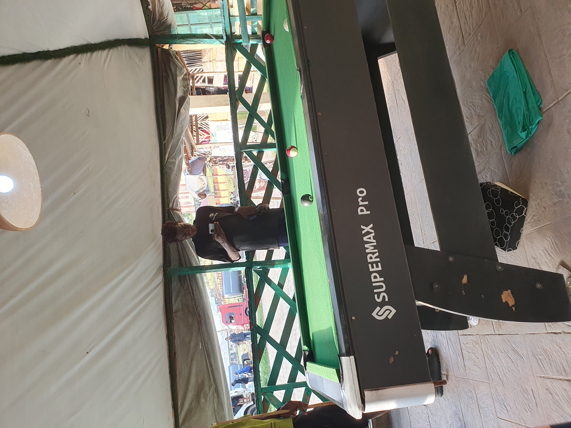 In Lokoja, Nigeria, a billiard table, identified as a "SUPERMAX Pro," is the central focus of this image, situated outdoors under a white canopy. The green felt surface of the table is dotted with several billiard balls: a black eight ball, a red ball, and a striped ball with red and white markings. A white cue ball is also visible. A young man, wearing a black t-shirt and shorts, stands to the right of the table, engrossed in his mobile phone. To the left, another person, partially visible, stands with a billiard cue. In the background, beyond a green wooden railing, traffic and buildings suggest an urban environment. The ground beneath the table is paved with textured tiles, with a folded green cloth lying on the ground near the table's legs.