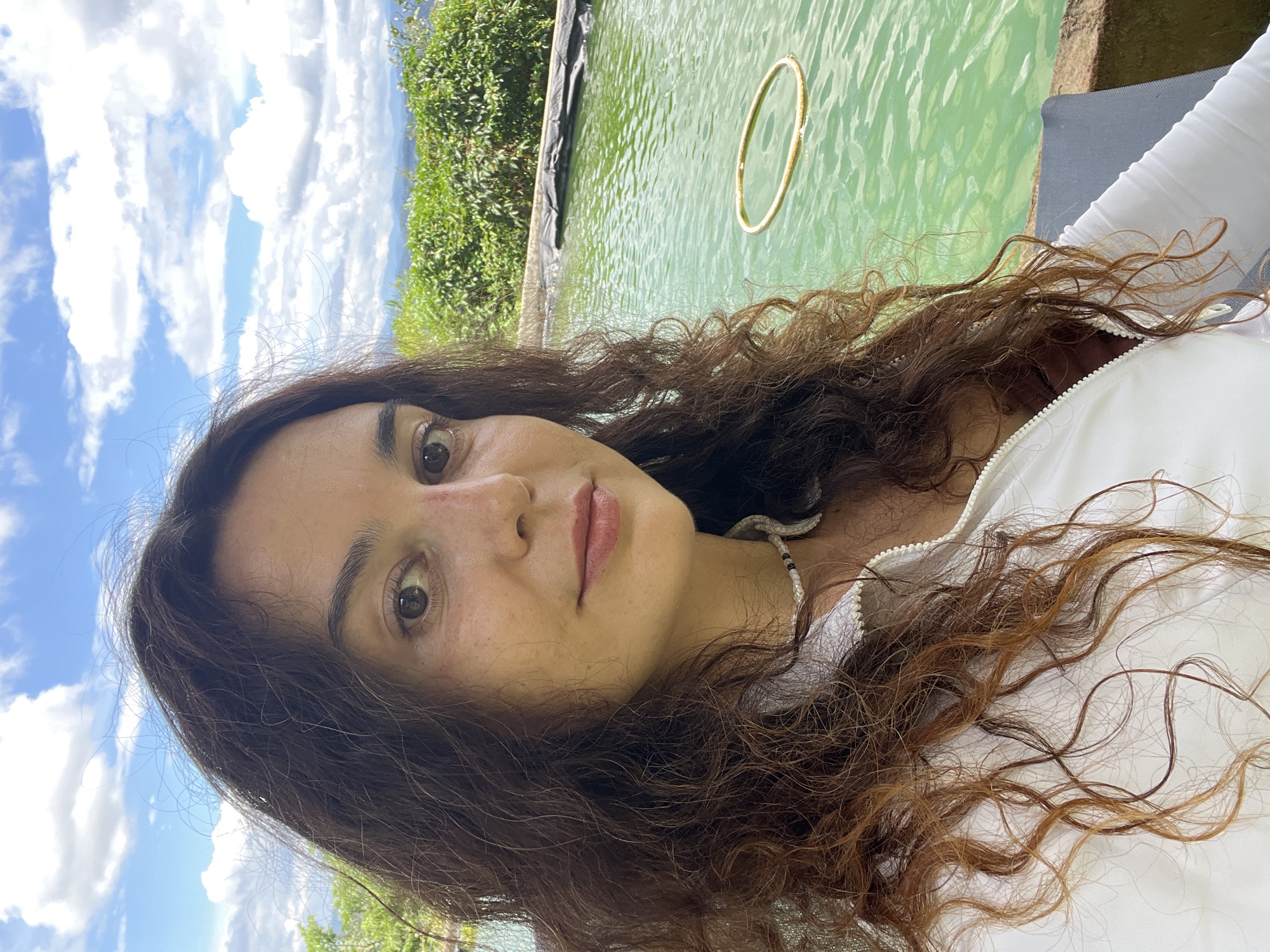 A woman with long, dark, curly hair is taking a selfie outdoors. She is wearing a white zip-up jacket over a brown top and a beaded necklace. Her left eye is visible, along with part of her nose and mouth. She appears to be outdoors, with a pool of green water behind her. There is a golden ring floating in the water. Lush green foliage and a cloudy blue sky are visible in the background. The scene suggests a resort or a scenic natural setting, possibly in the rural outskirts of Sơn Điền, Vietnam, given the natural vegetation and open sky.