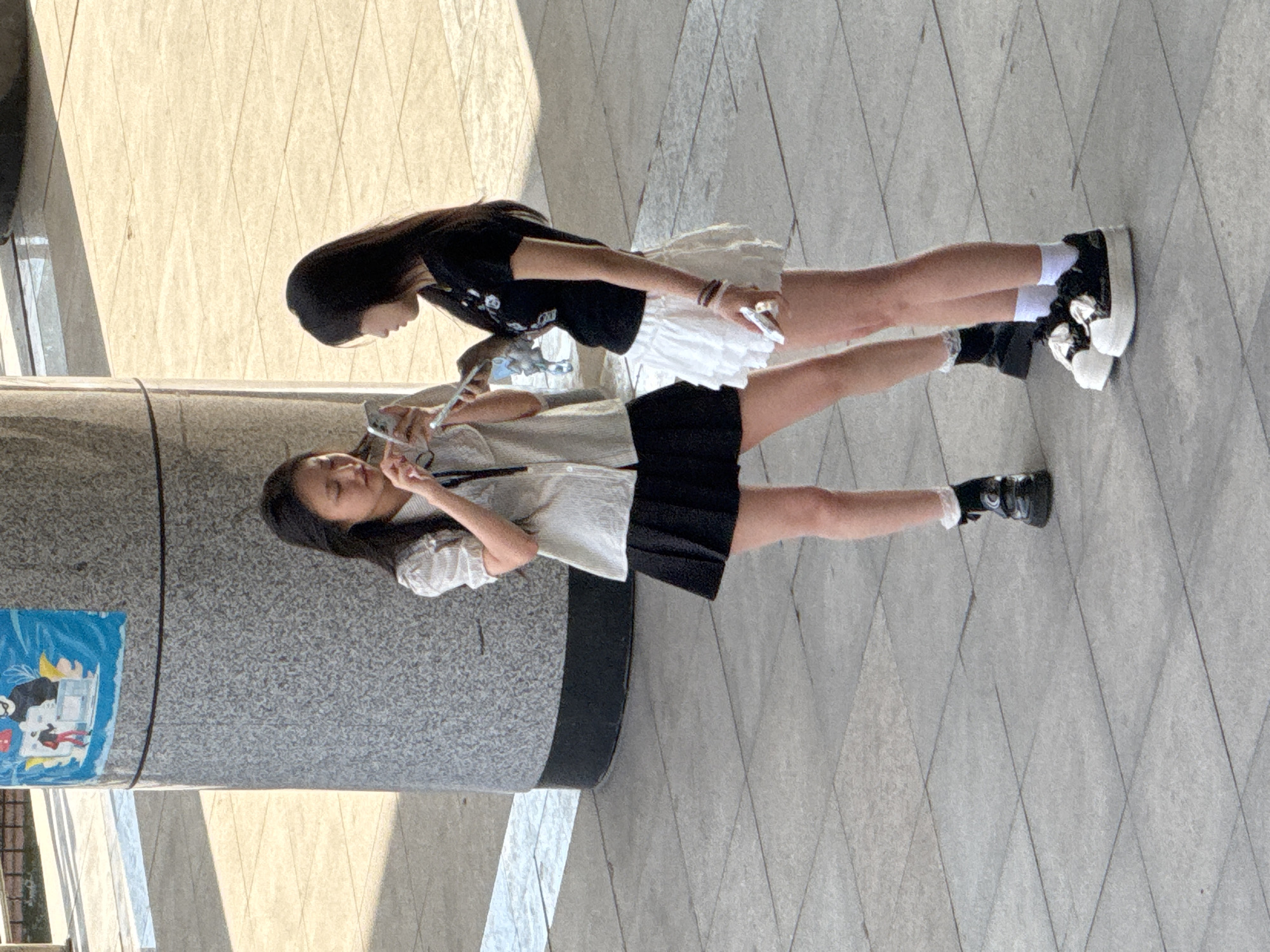 Two young women are standing on a paved outdoor area, possibly a plaza, in the city of Guta, China. The woman on the left is wearing a light-colored short-sleeved shirt and a black pleated skirt, black socks with white frills, and black shoes. She is holding a smartphone in her hands, looking down at it. The woman on the right is wearing a black t-shirt and a white ruffled skirt, white socks, and black and white platform sneakers. She is also holding a smartphone, looking down at it. The two women appear to be interacting with their phones. Behind them, a large, gray, cylindrical pillar with a black base stands prominently. A poster with blue, white, and red graphics, depicting cartoon characters, is affixed to the pillar. The ground is tiled with square concrete slabs. Sunlight casts shadows across the ground, indicating it is daytime.