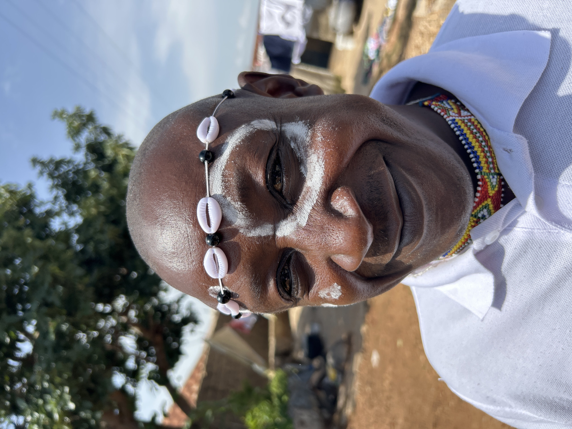 This is a close-up, vertically oriented, eye-level shot of a dark-skinned person wearing traditional adornments. The individual has white face paint applied in a decorative pattern around their eyes and on their forehead. A string of black beads interspersed with white cowrie shells is worn as a headband. They are also wearing a colorful beaded necklace that is visible beneath the collar of a white collared shirt.
The background suggests an outdoor setting in a dry, possibly rural or semi-urban area, with some vegetation and buildings visible. The lighting indicates it is daytime, likely bright sunlight. The overall impression is one of cultural or spiritual significance, given the face paint and traditional jewelry. There is no visible text.