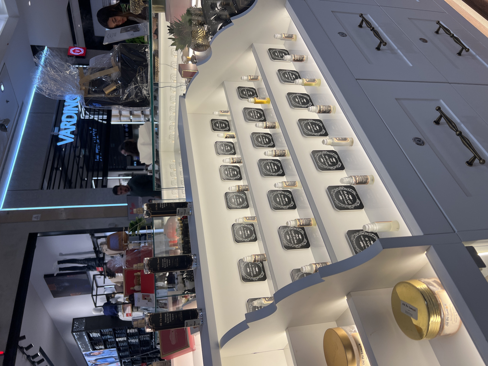 The image depicts an indoor commercial retail environment, specifically within a shopping mall or similar center in Kiryat Bialik, Israel. In the foreground, a multi-tiered white display cabinet showcases numerous small, clear glass rollerball bottles, each accompanied by a black square plaque with white Hebrew text indicating product names or descriptions. Larger clear jars with gold lids, labeled "Oil De La Mer," are positioned on the lowest visible shelf. The base of this cabinet features light grey drawers equipped with dark, ornate pull handles and keyholes.
Behind this main display, a glass counter holds several larger cylindrical clear glass bottles, branded "Oil De La Mer - PERFUME BODY MIST INTENSE." A black paper gift bag, encased in clear plastic wrap and adorned with a gold ribbon, rests on this counter to the right. Decorative gold pineapple-shaped objects are also present on the counter.
In the background to the right, a store named "VARDINGO" is visible, illuminated by a prominent neon blue sign. A female individual with dark hair is seated at a counter within the "VARDINGO" store, looking downwards at an object, likely a tablet or mobile phone. To the background left, another retail space, possibly a clothing store, is evident with mannequins, folded apparel on shelves, and digital screens displaying merchandise. A male individual in a dark green shirt is partially visible near the entrance of this store. The scene is illuminated by a combination of overhead track lighting, display case accent lighting, and neon signs, consistent with a contemporary retail setting.